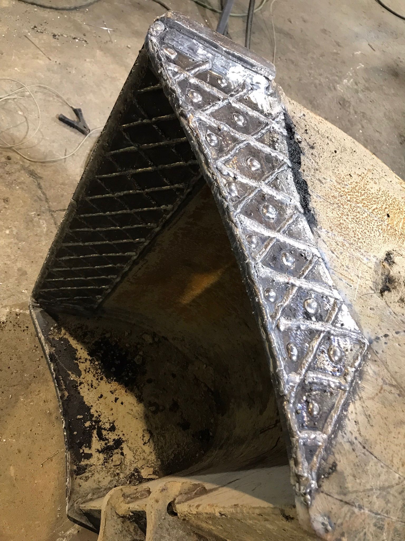 Silver, diamond-patterned metal duct on sandy ground, open at one end, with dark debris inside.