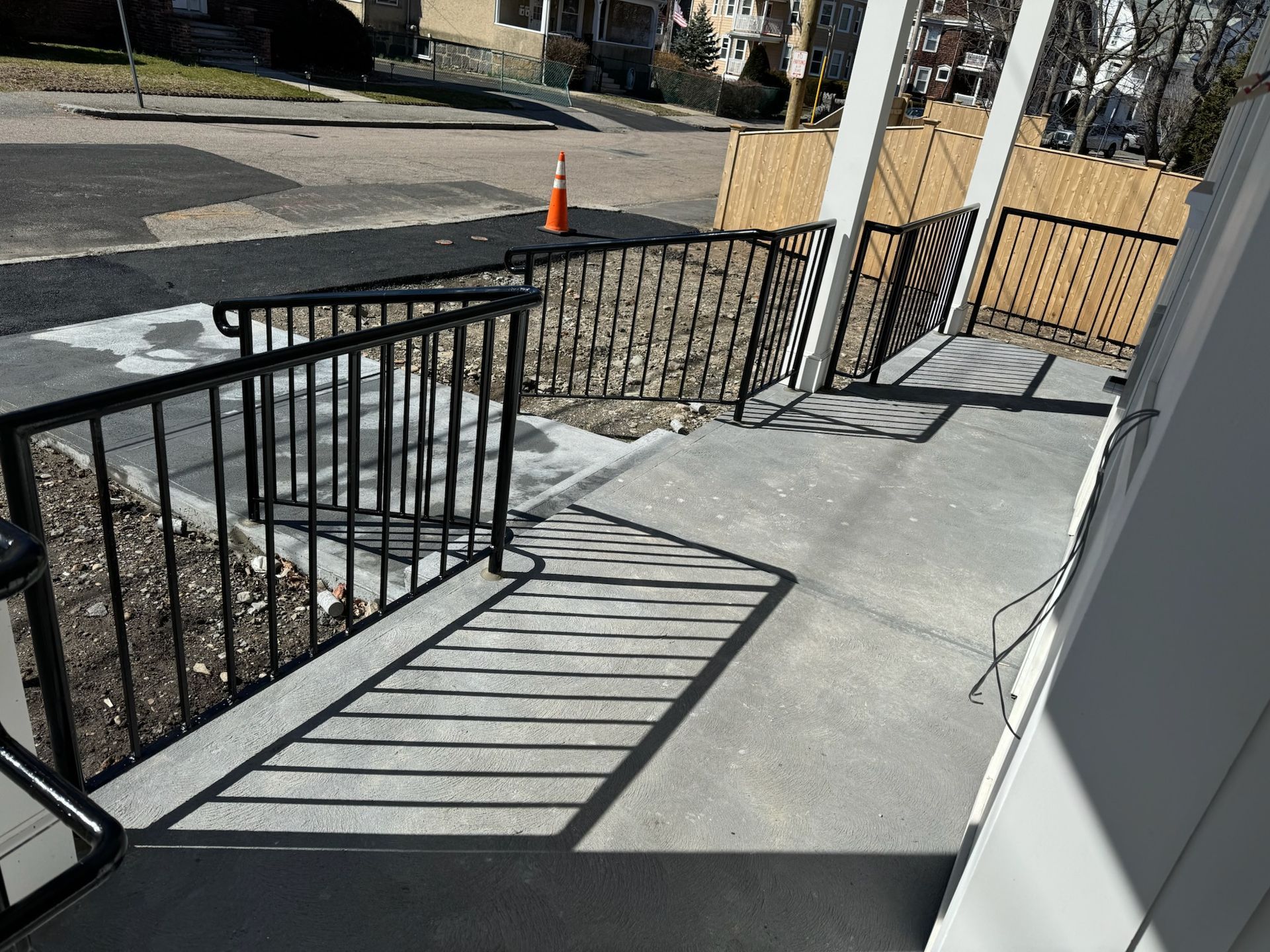 Ramps with black railings leading to a sidewalk, with an orange cone and fenced area in the background