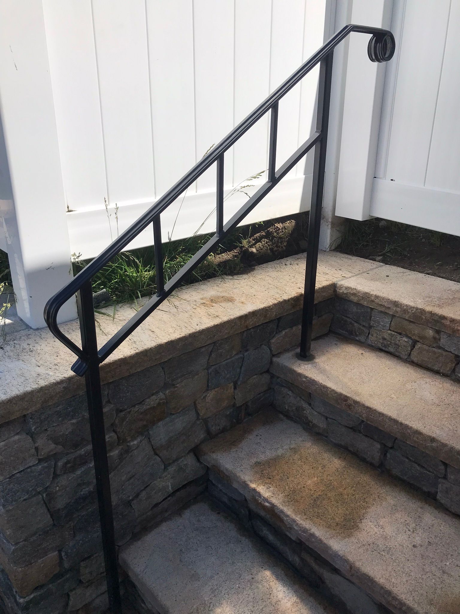 Black metal handrail beside stone steps and retaining wall next to a white fence.