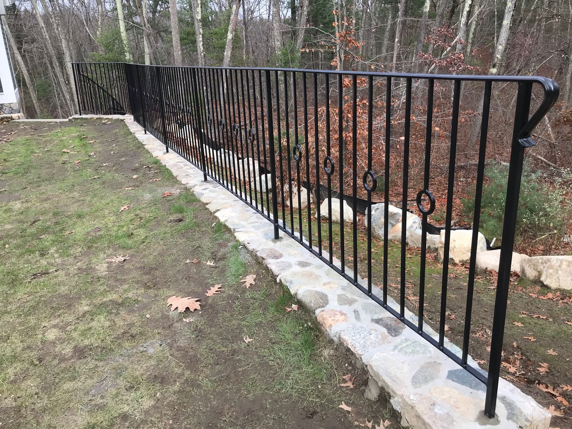 Black metal fence along a stone border beside a grassy yard and leafless woods.