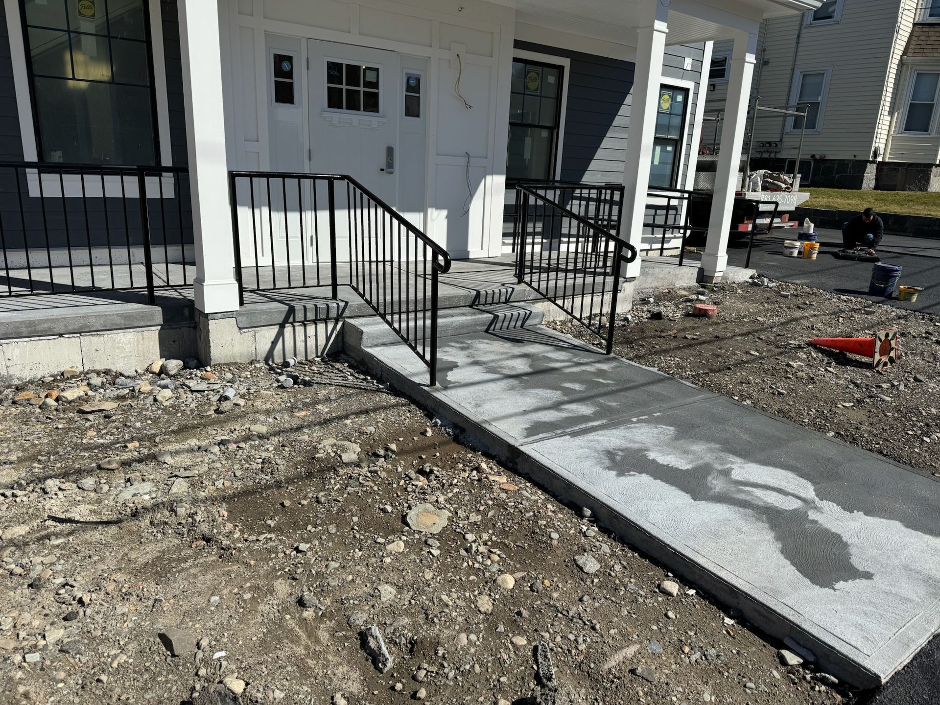 Front porch with concrete ramp and black railings beside a white house, on a dirt construction lot
