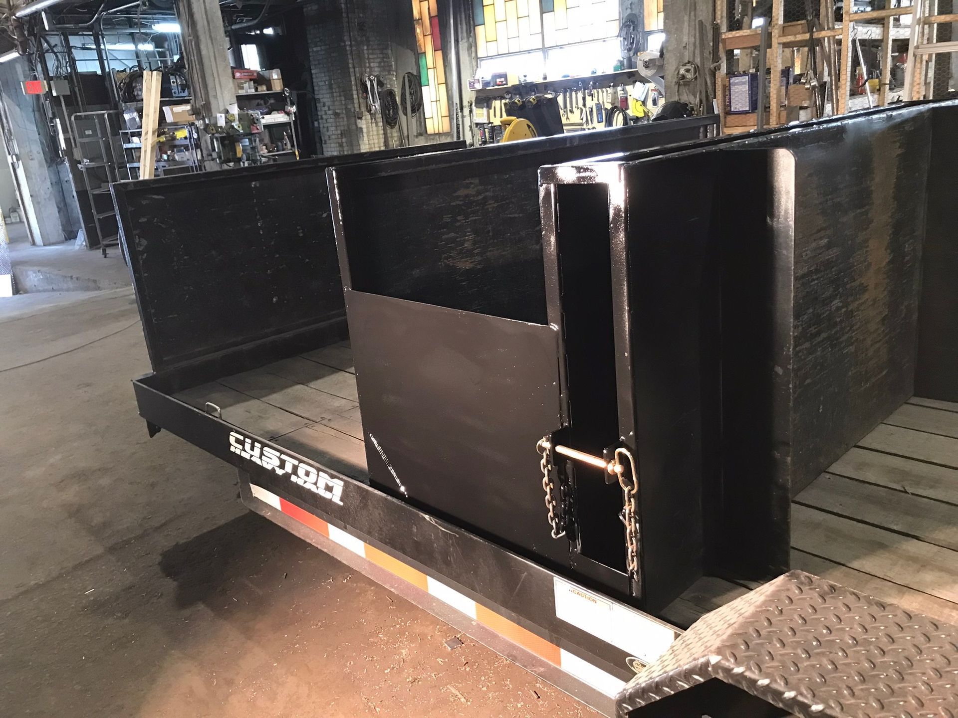 Black metal utility trailer with open side gate parked indoors under bright lights