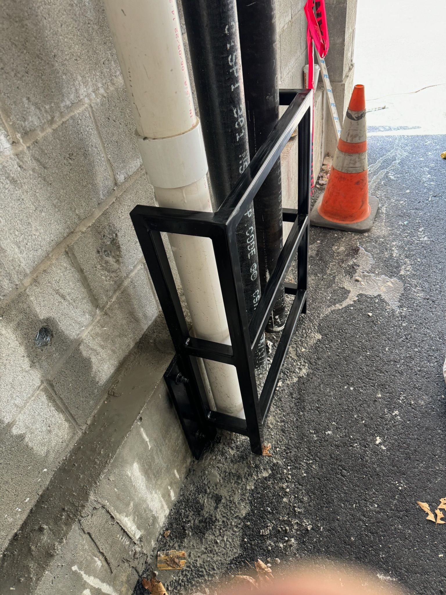 Black metal railing around white pipes beside a concrete wall, with an orange traffic cone on wet pavement.