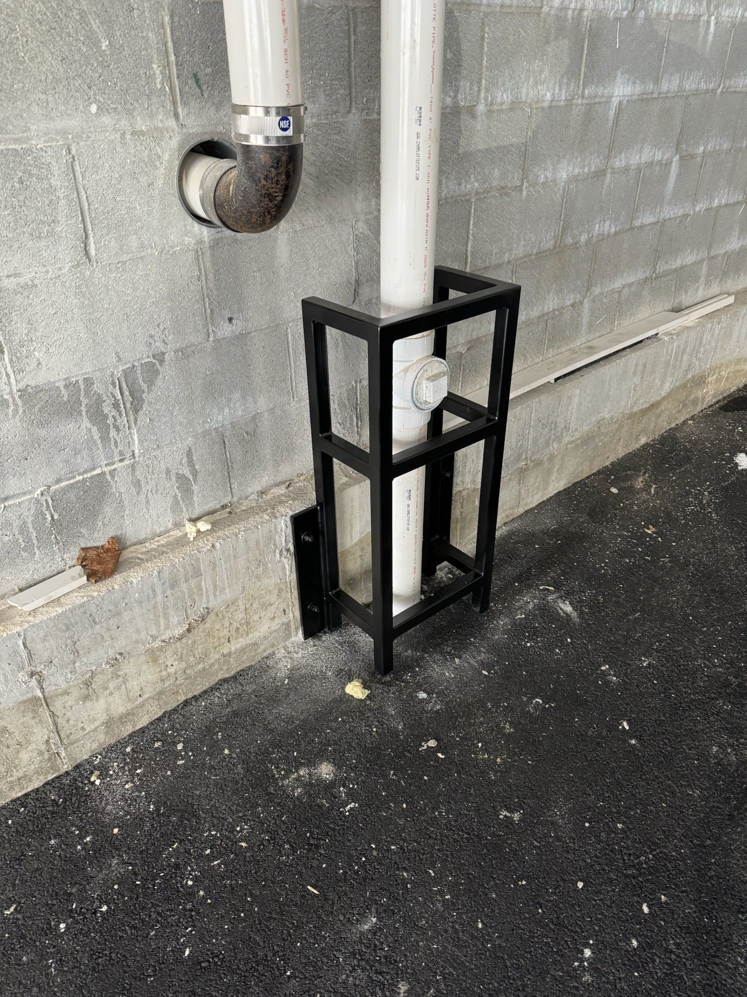 Black metal pipe guard around a white downspout against a concrete wall on asphalt ground