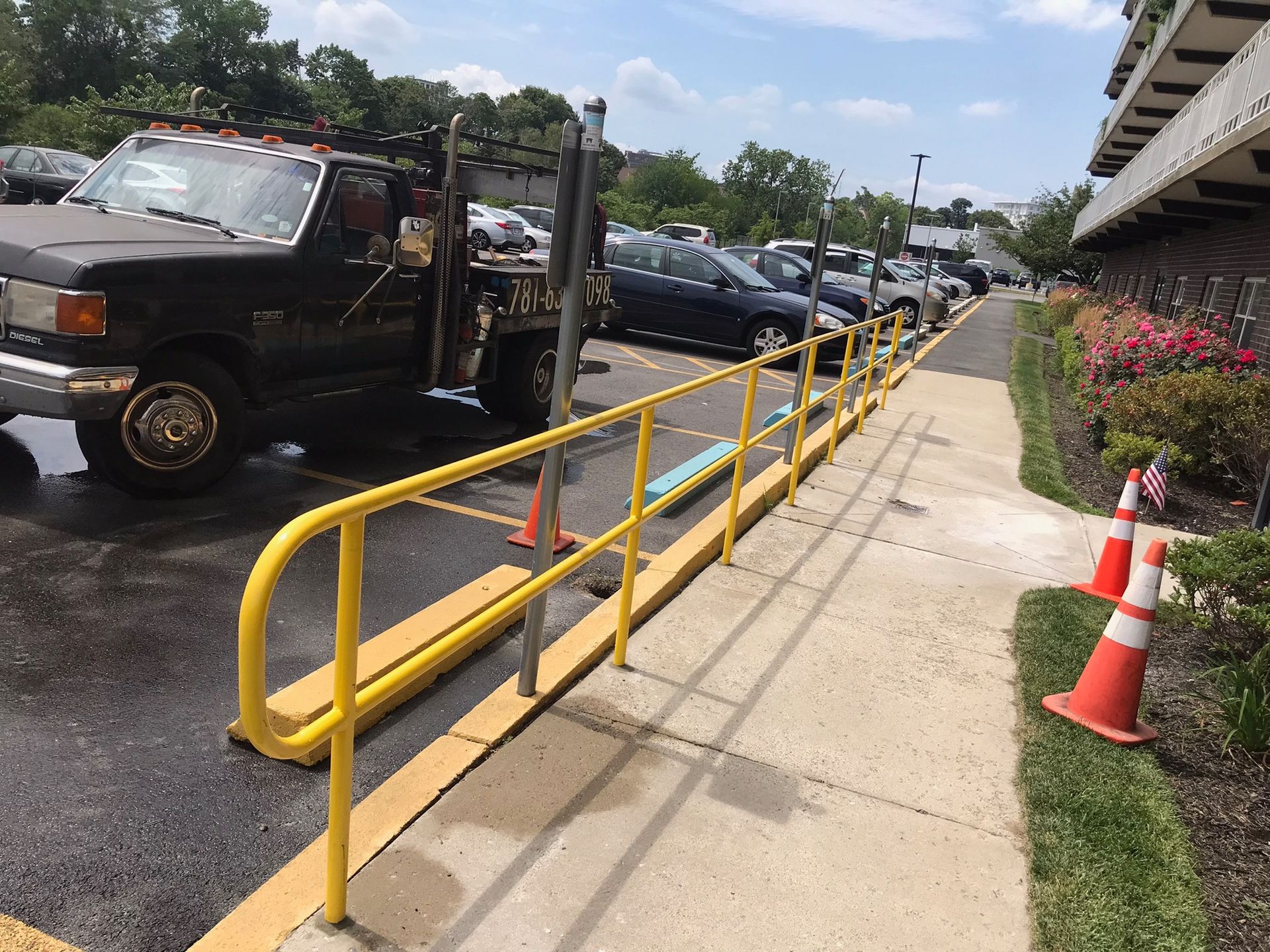 Yellow ramp rail along a sidewalk beside parked vehicles and orange cones near a building