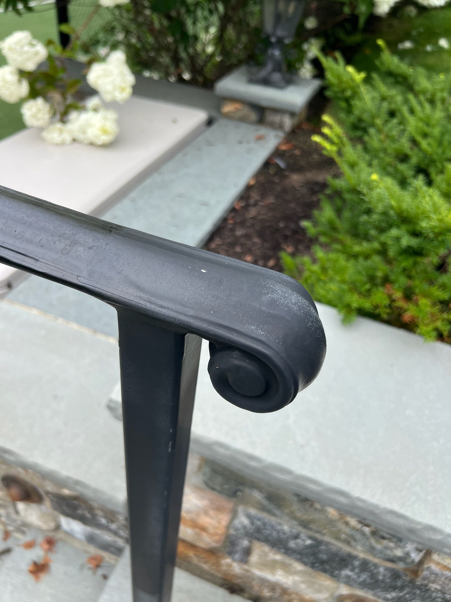 Black metal stair railing with a rounded end beside a garden and stone steps.