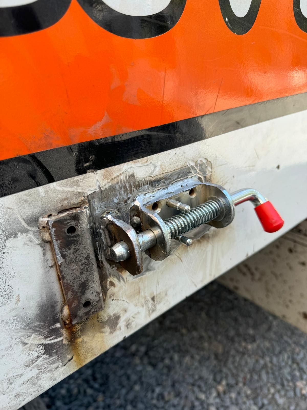 Close-up of a metal latch with a red handle on a dirty orange and white vehicle panel