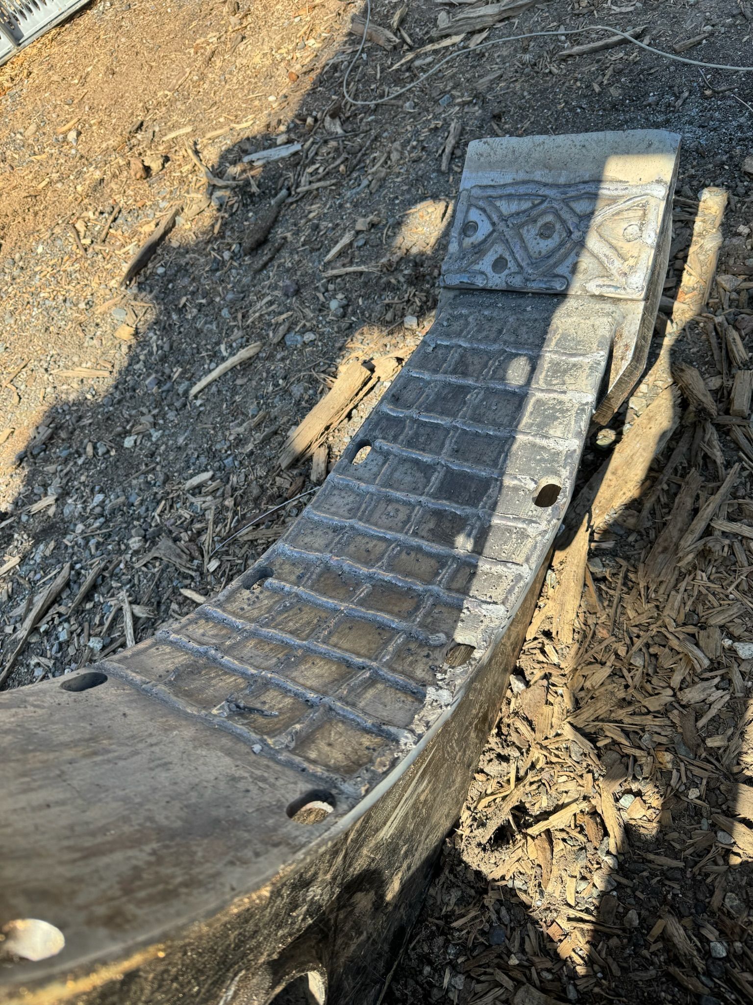 Broken black plastic curved ramp edge on a gravel path, with a shadow cast across it.