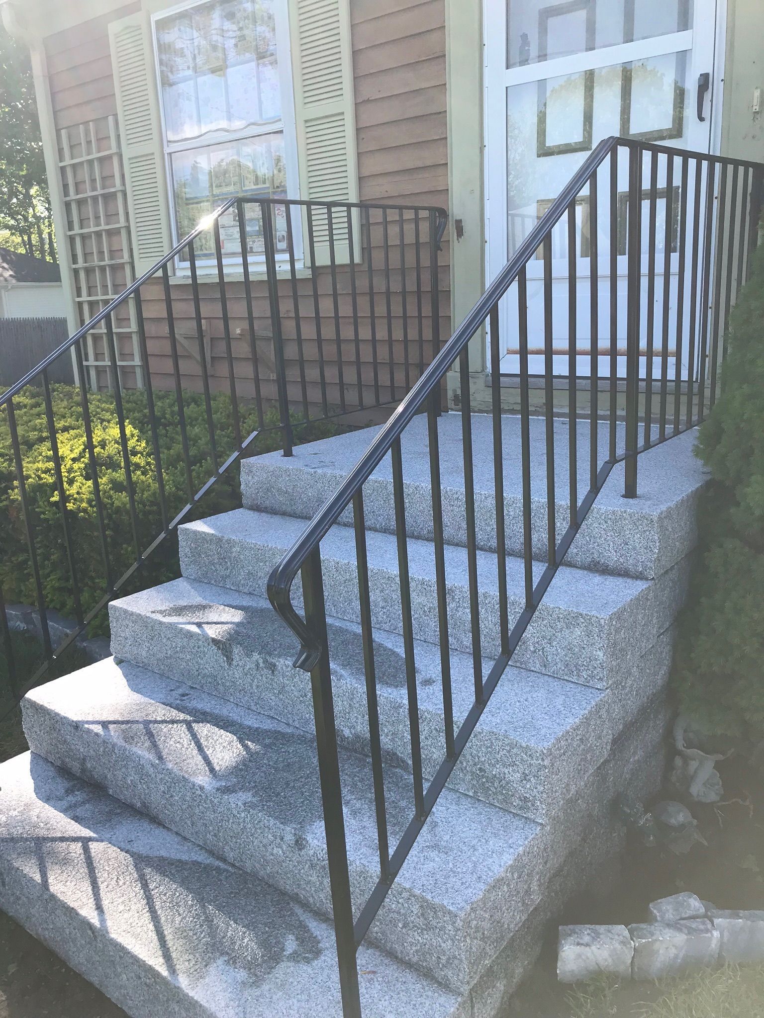 Front steps with black railings leading to a house entrance, beside a window and landscaped shrubs.