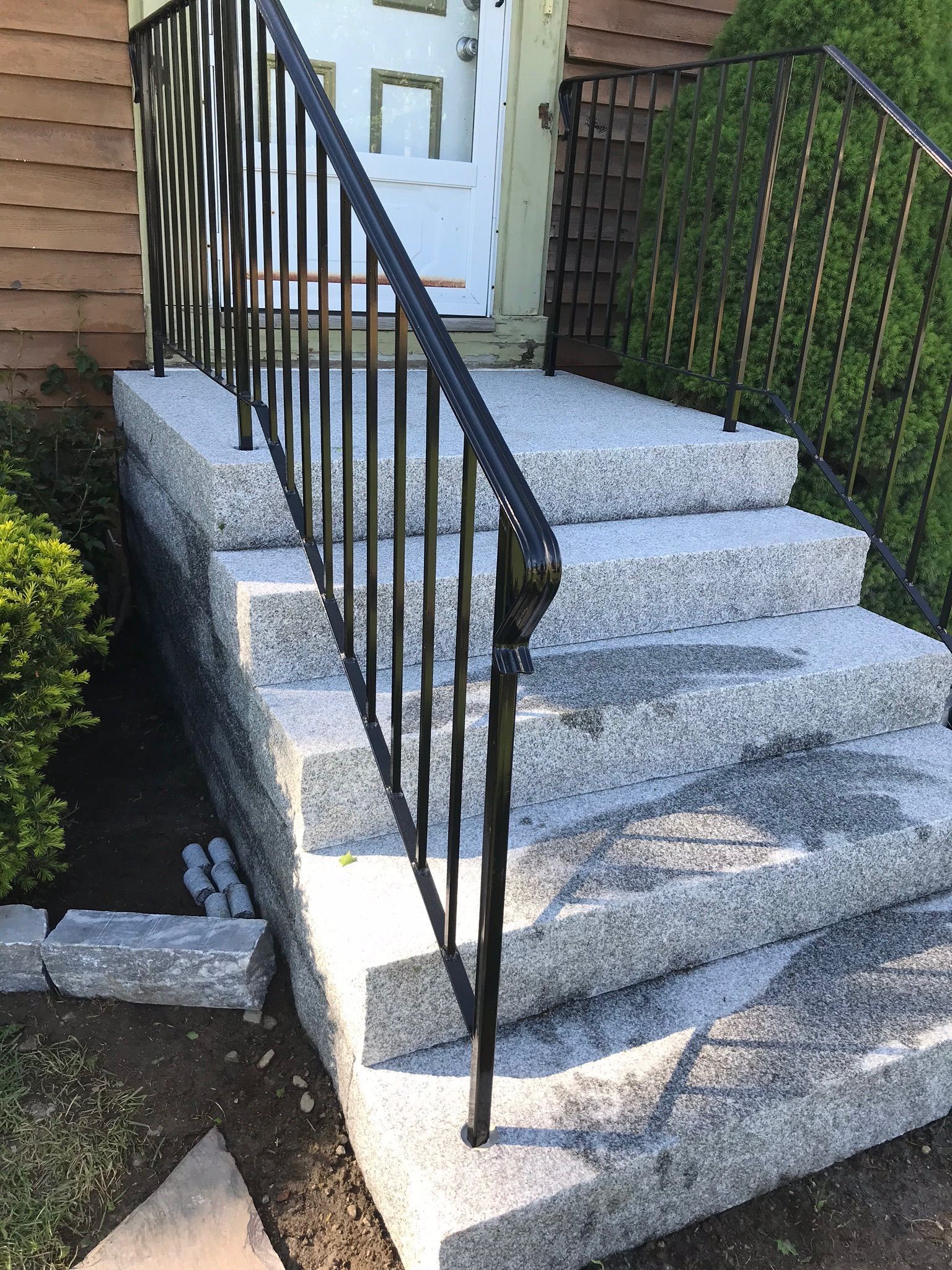 Front steps with black metal railing leading to a white door, bordered by shrubs and brick siding.