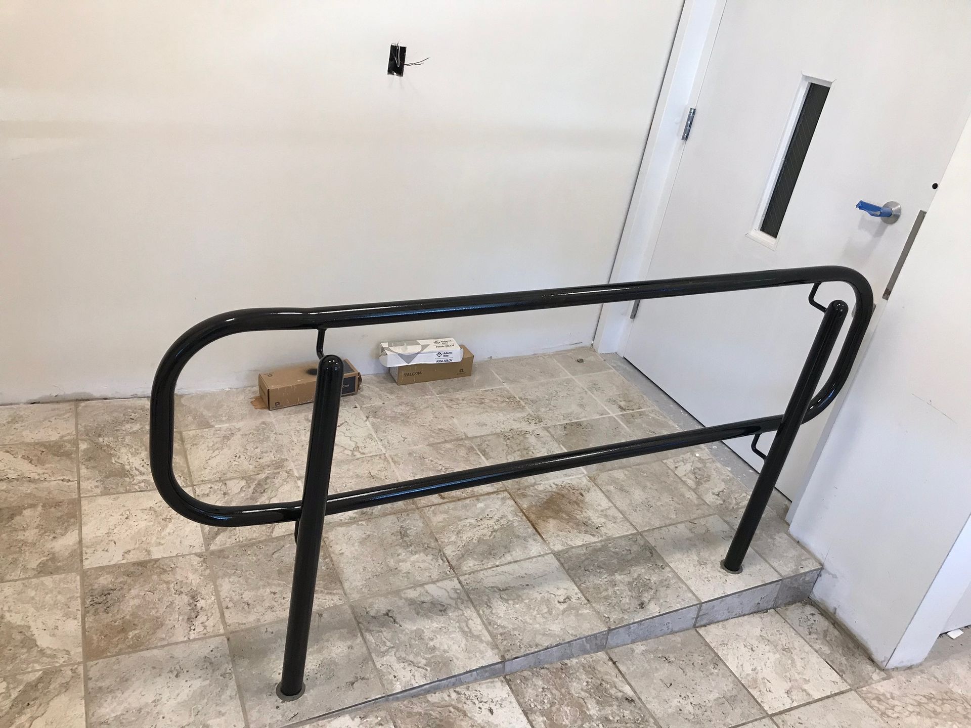 Black metal handrail in a tiled indoor space near a white door and wall-mounted fixture.