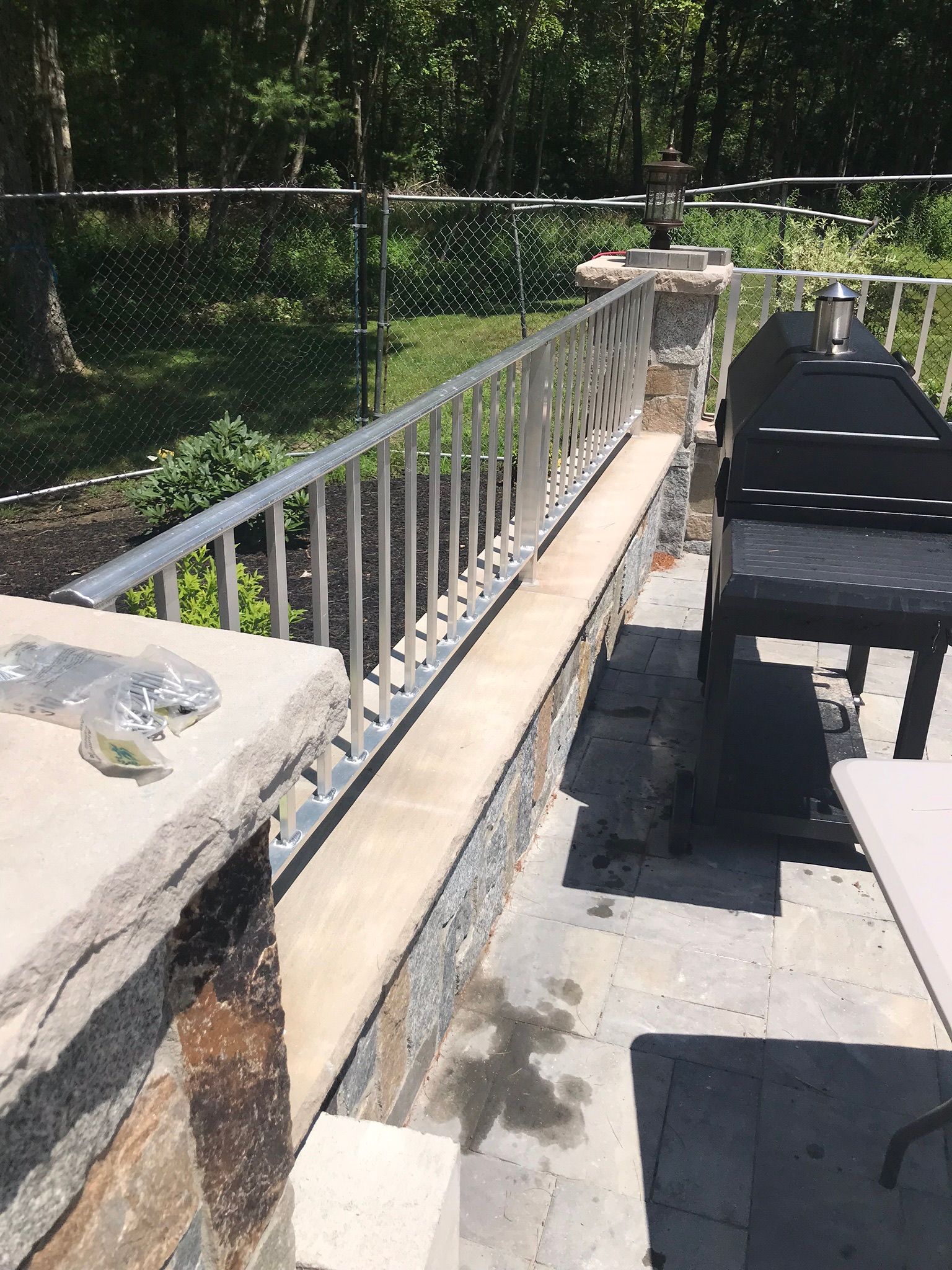 Outdoor patio with metal railing beside a grill, stone surface, and greenery beyond