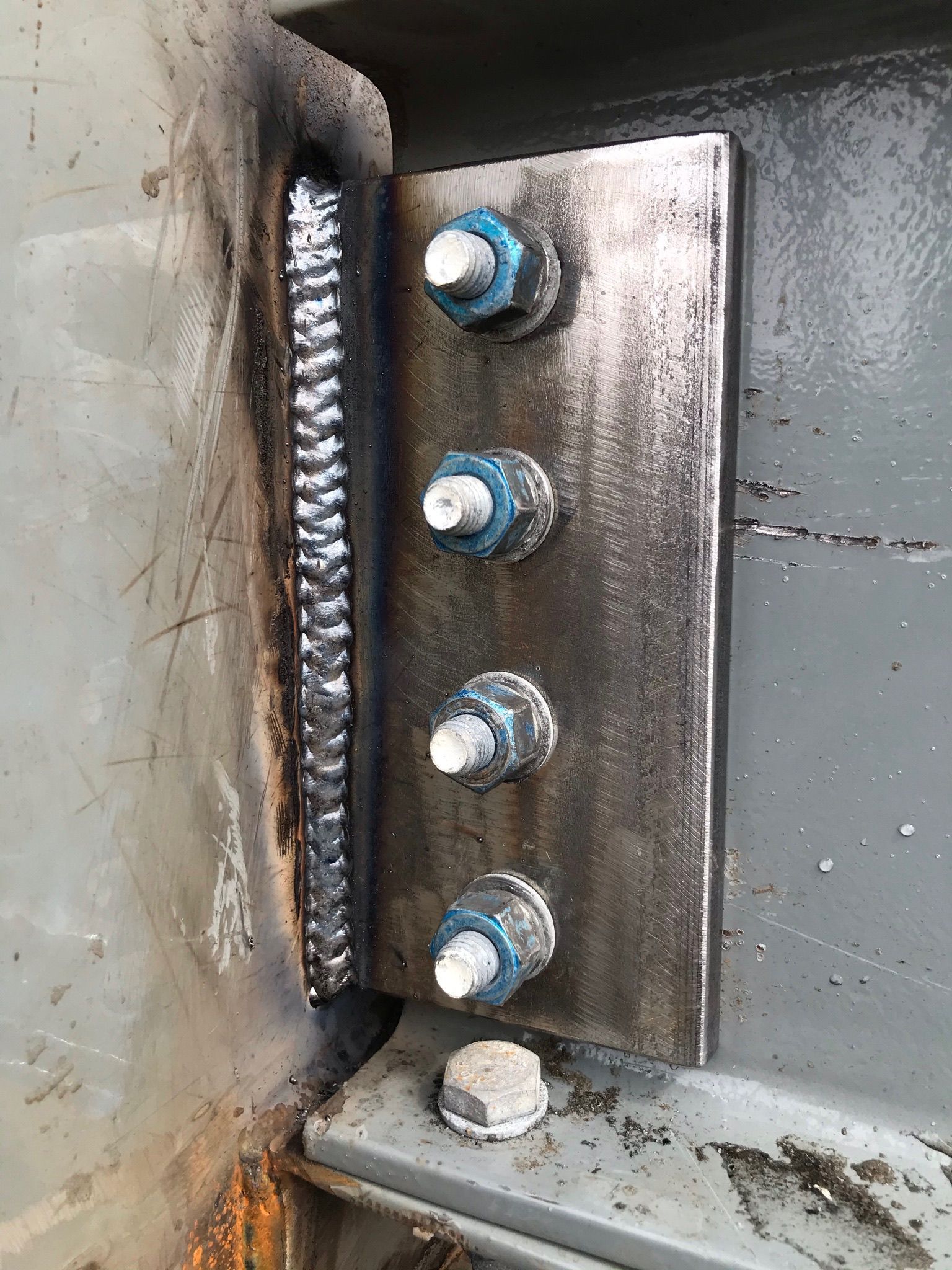 Close-up of a rusted metal hinge plate with four bolts on a gray industrial surface.