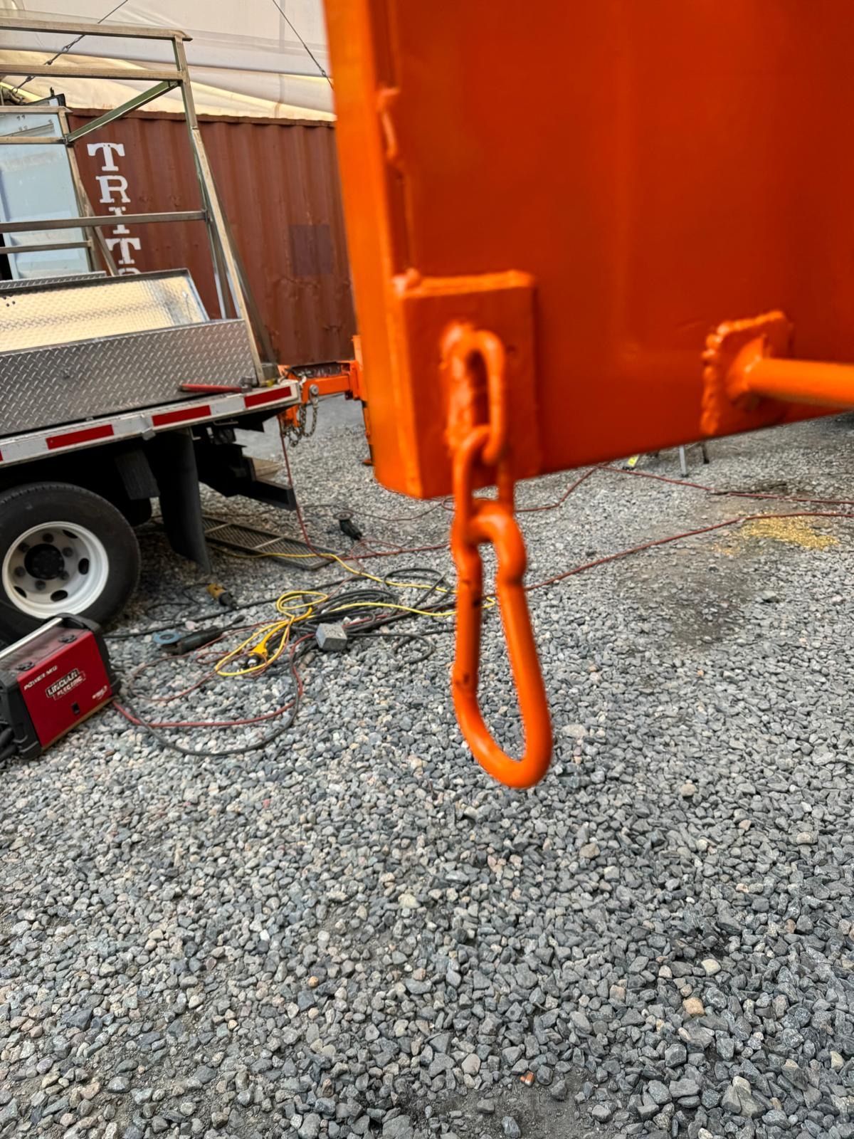 Orange metal hook hanging from a large orange container on a gravel lot near a truck.