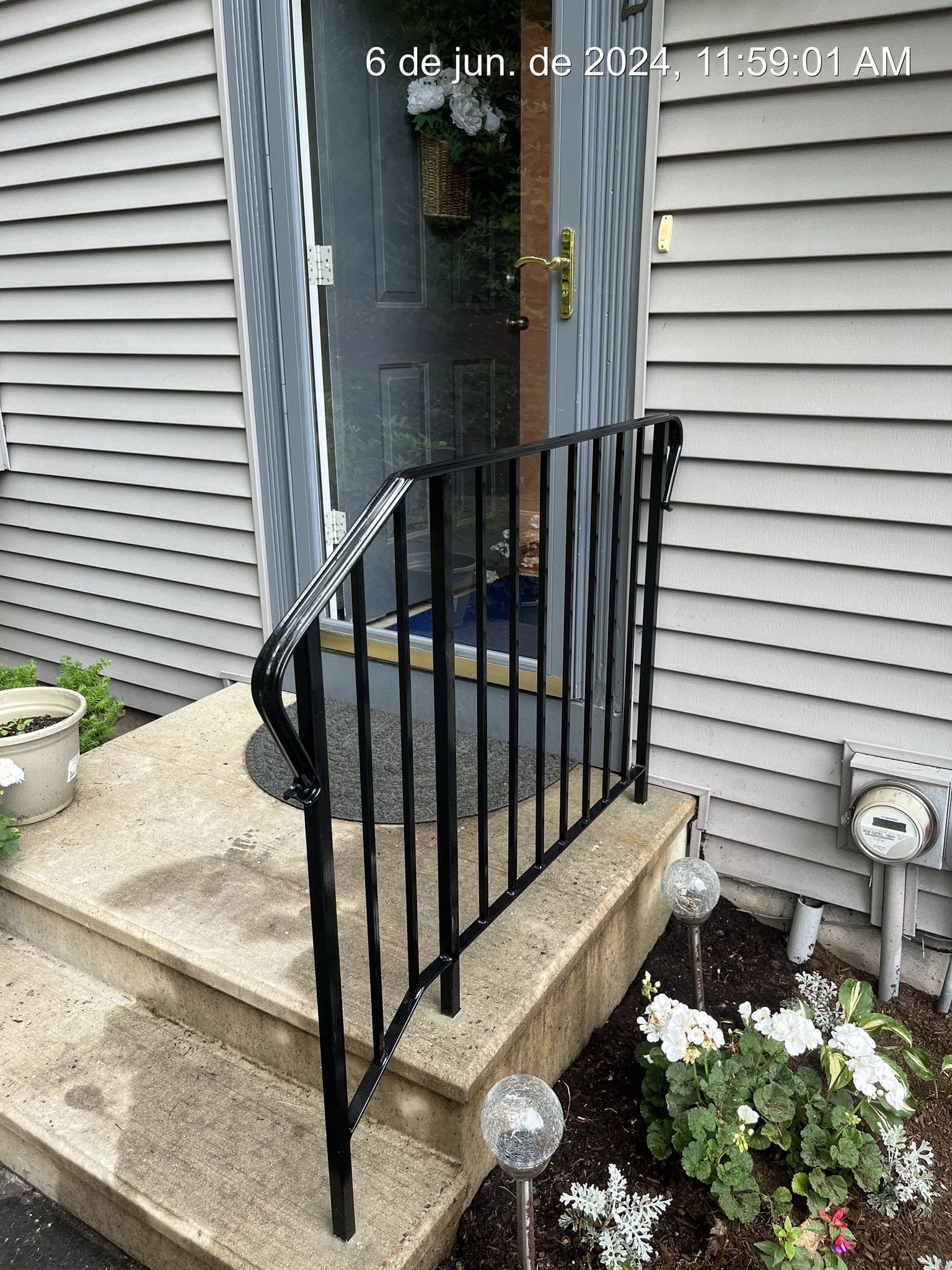 Front steps with black railing leading to a gray house door beside white siding and flower beds