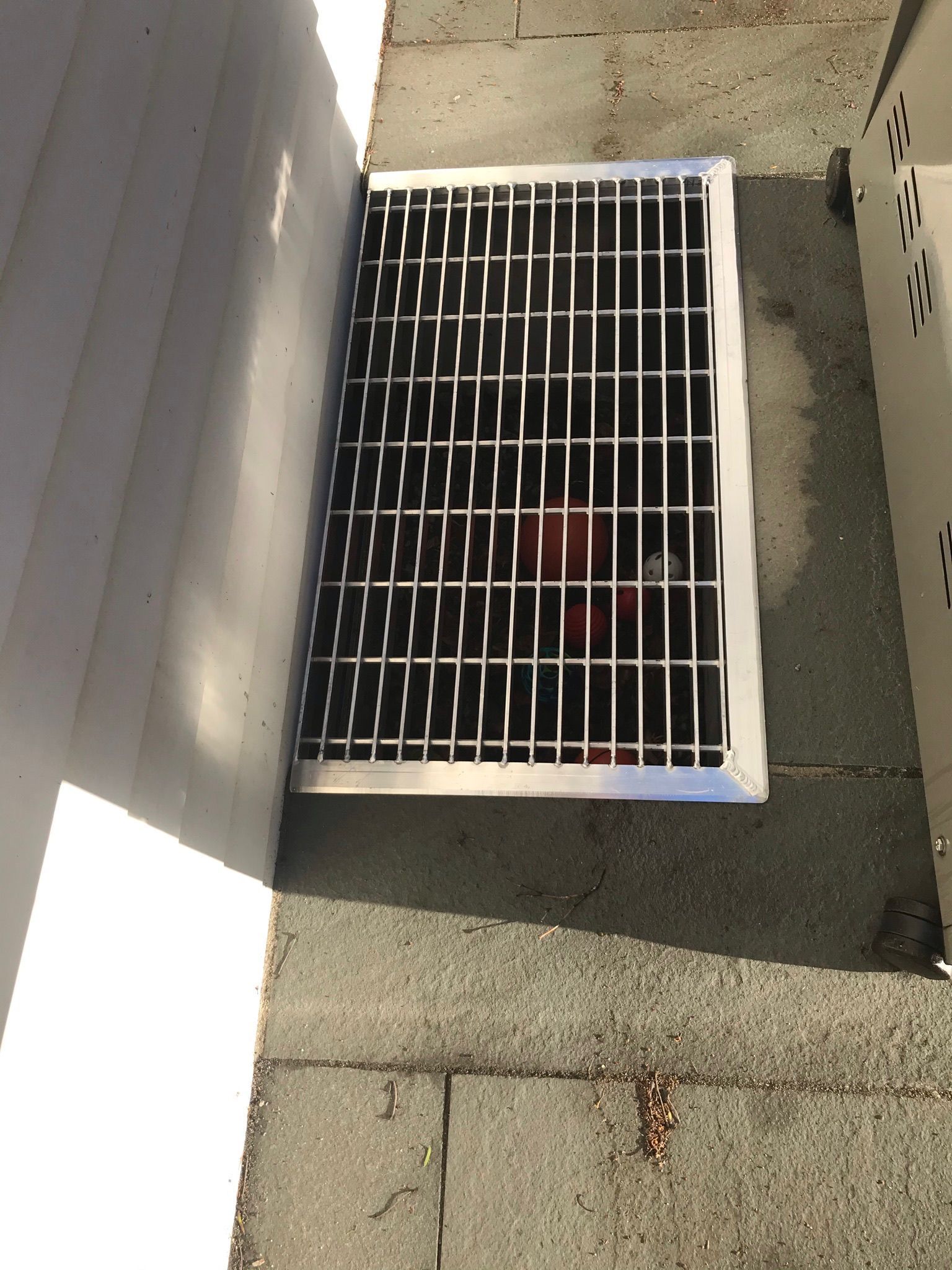 White metal vent grille beside a wall on a concrete surface, with a red object visible underneath.