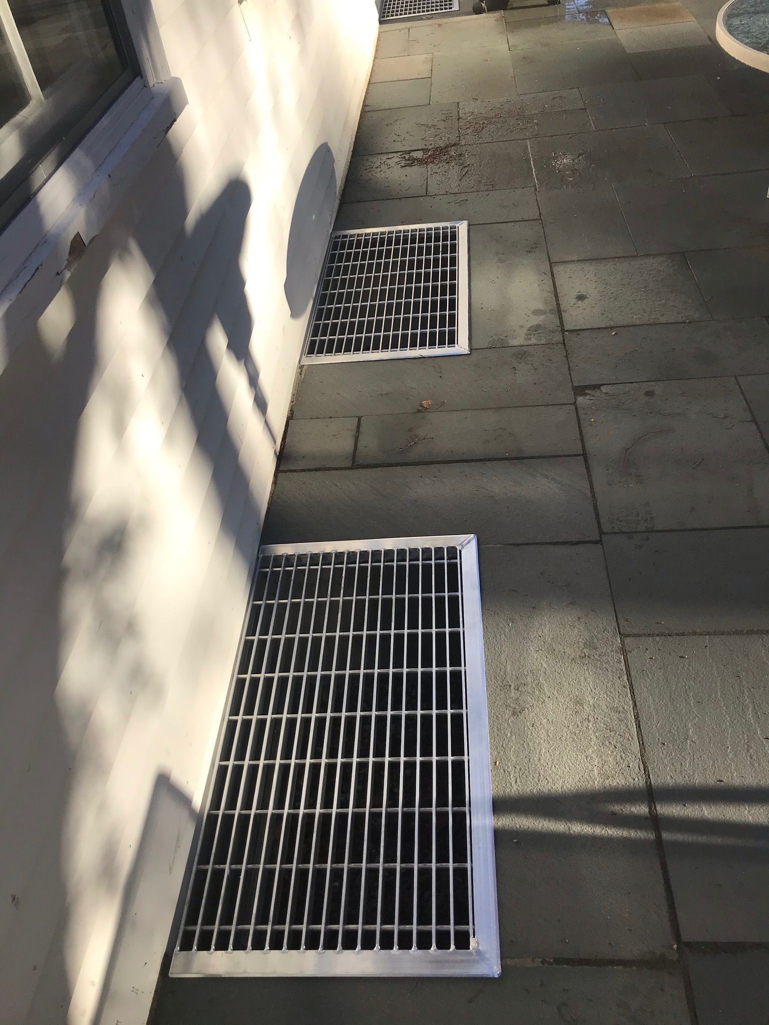 Sidewalk with two white metal grates beside a wall, sunlight casting shadows.