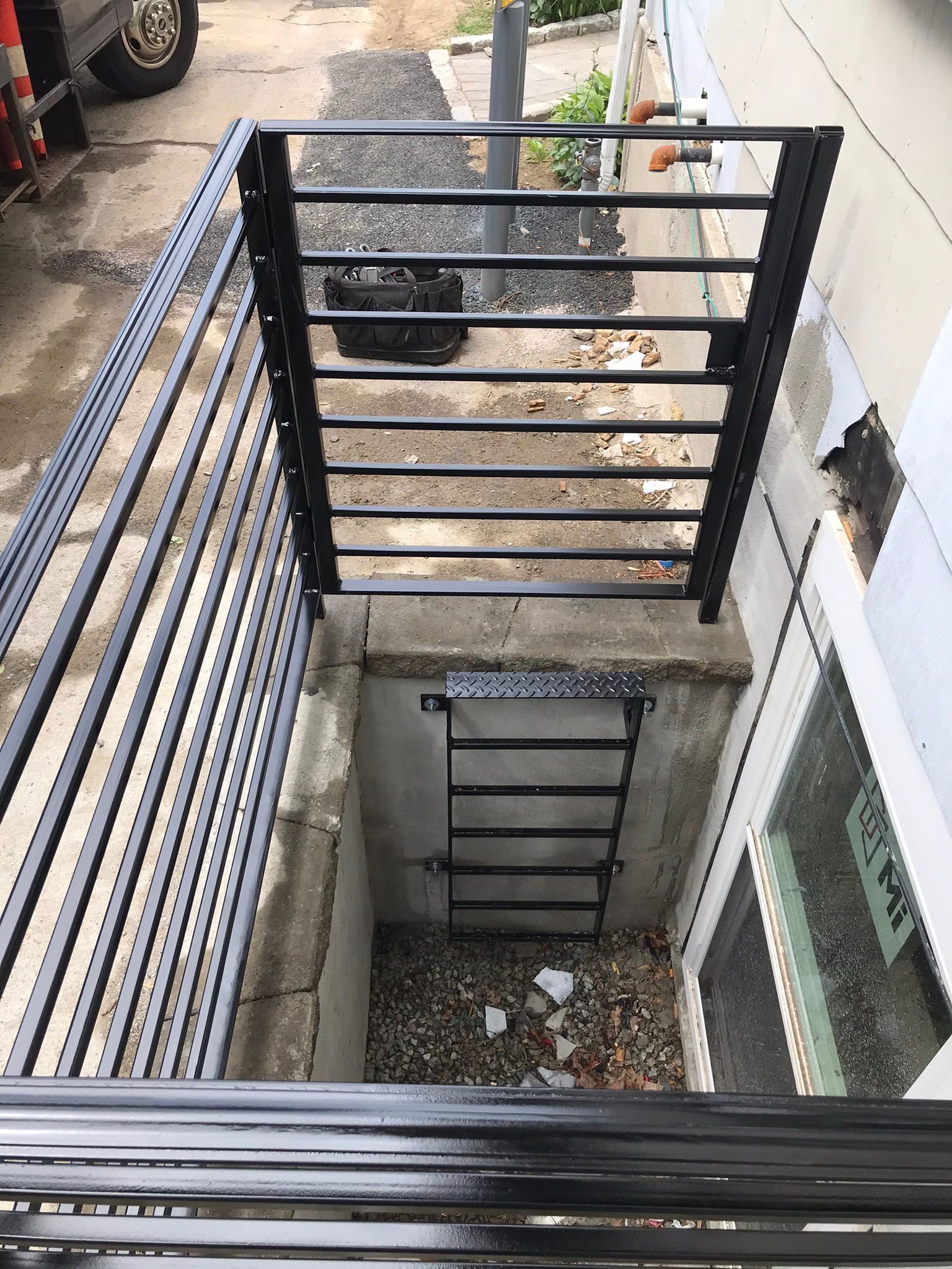 Black metal ladder descending into a narrow concrete utility shaft beside a building.