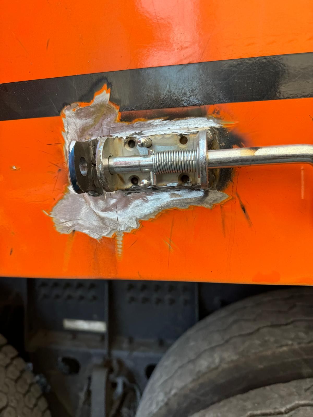 Close-up of a welded metal rod on an orange machine panel with a black stripe.