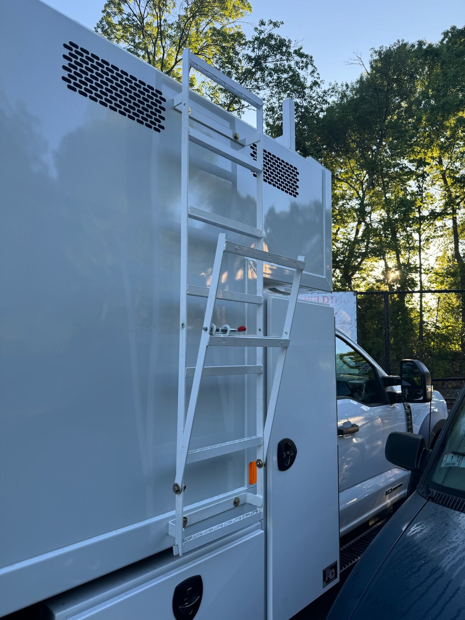 White utility truck with rear ladder rack parked outdoors beside a tree and another vehicle.