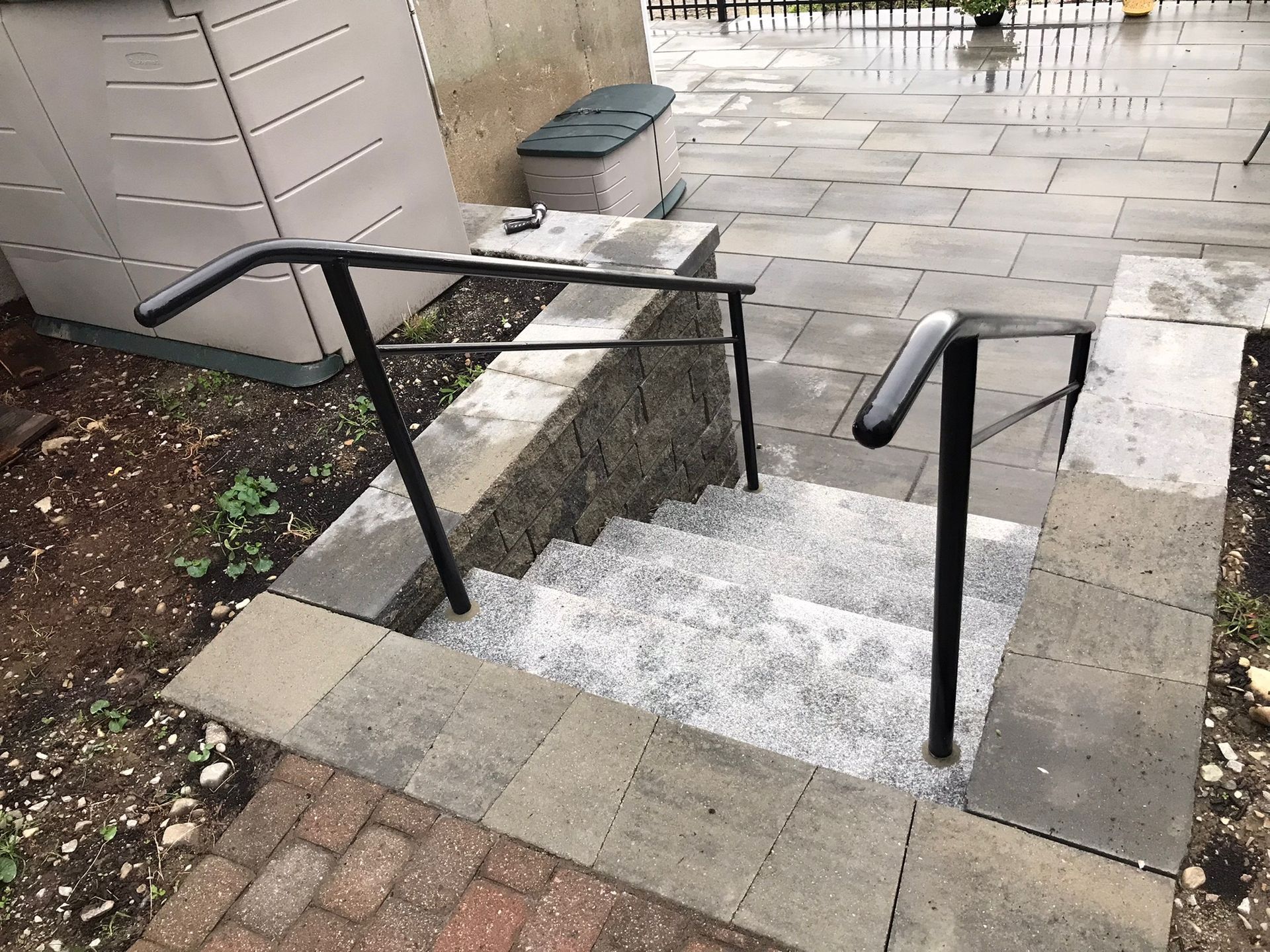 Steep outdoor stone steps with black handrails, bordered by brick and paving stones, after rain.