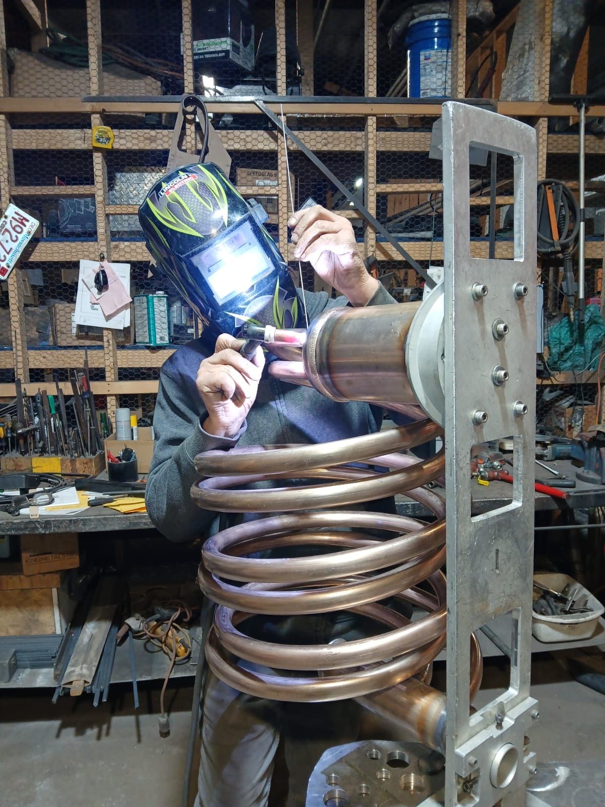 Person welding a large copper coil in a workshop, wearing a face shield and gloves