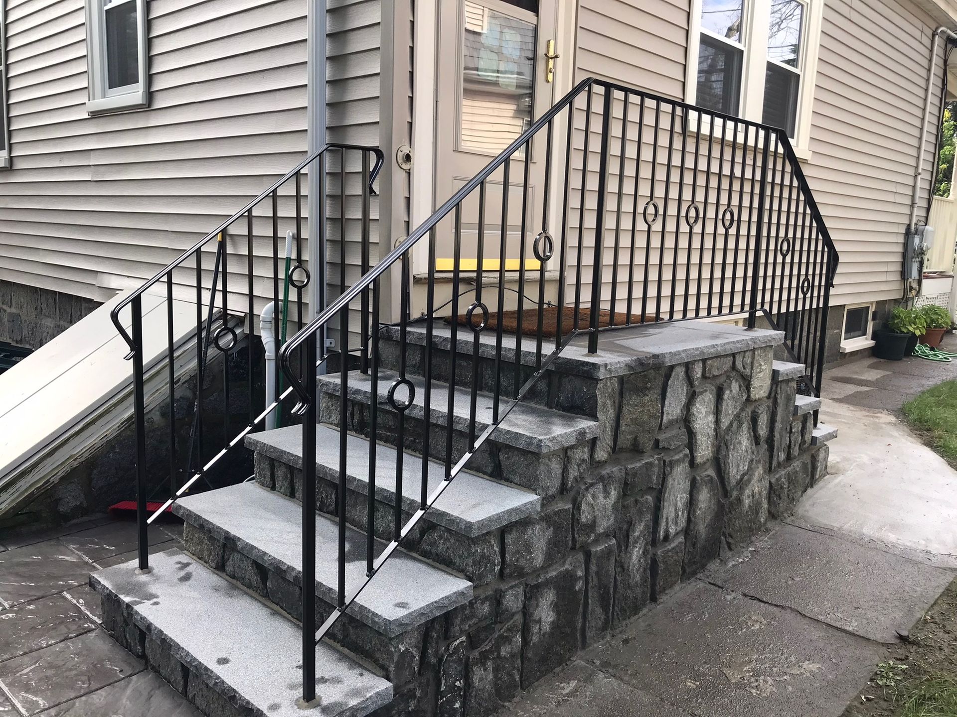 Front steps with stone retaining wall and black metal railing beside a house entrance