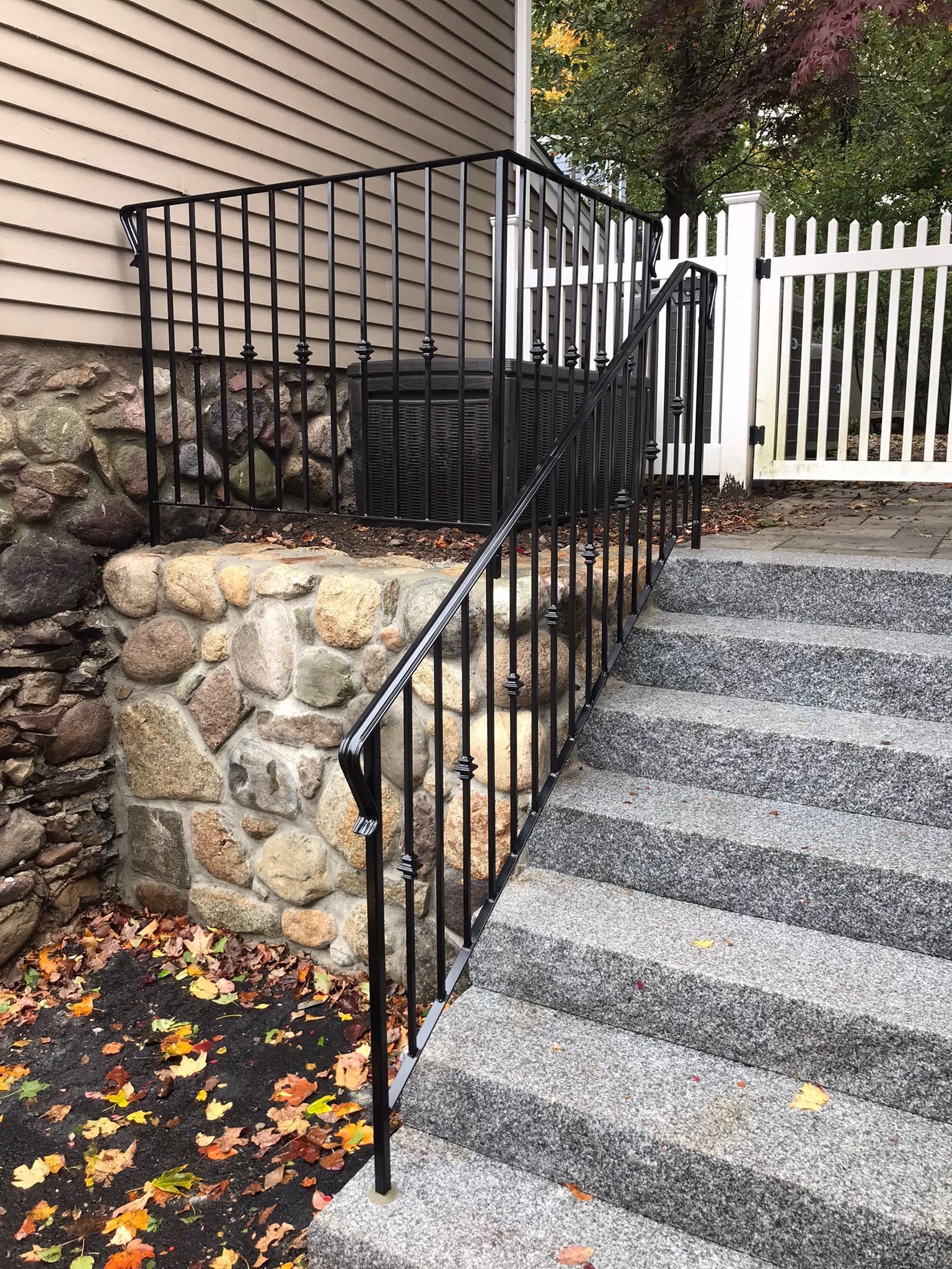 Outdoor stairway with black metal railing beside a stone retaining wall and white fence