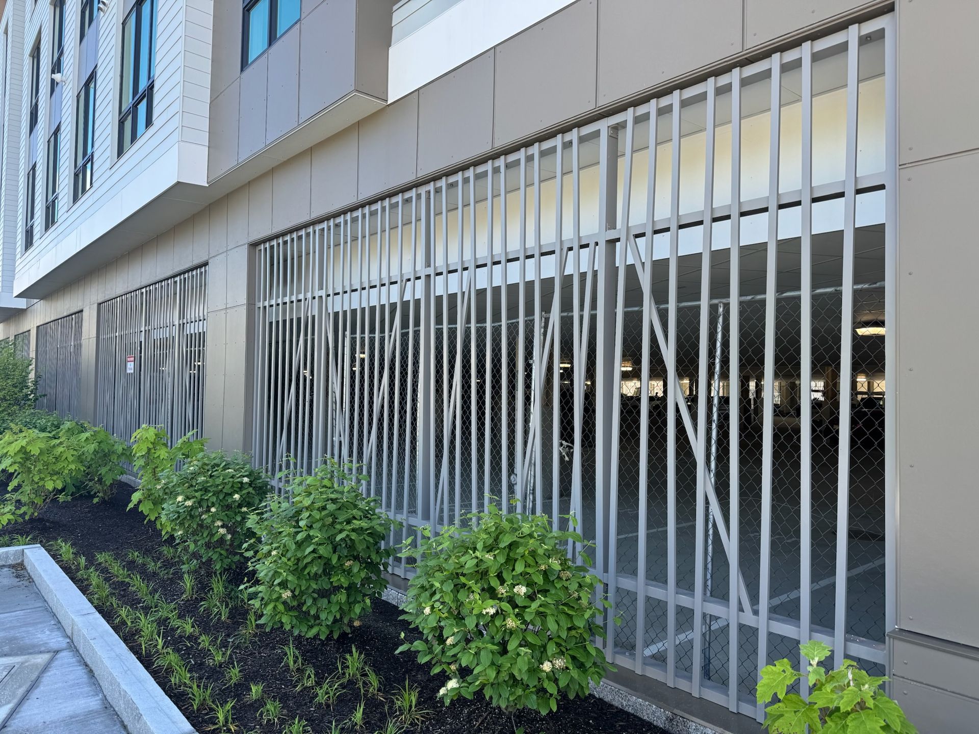 Modern building exterior with barred windows and green shrubs along the sidewalk