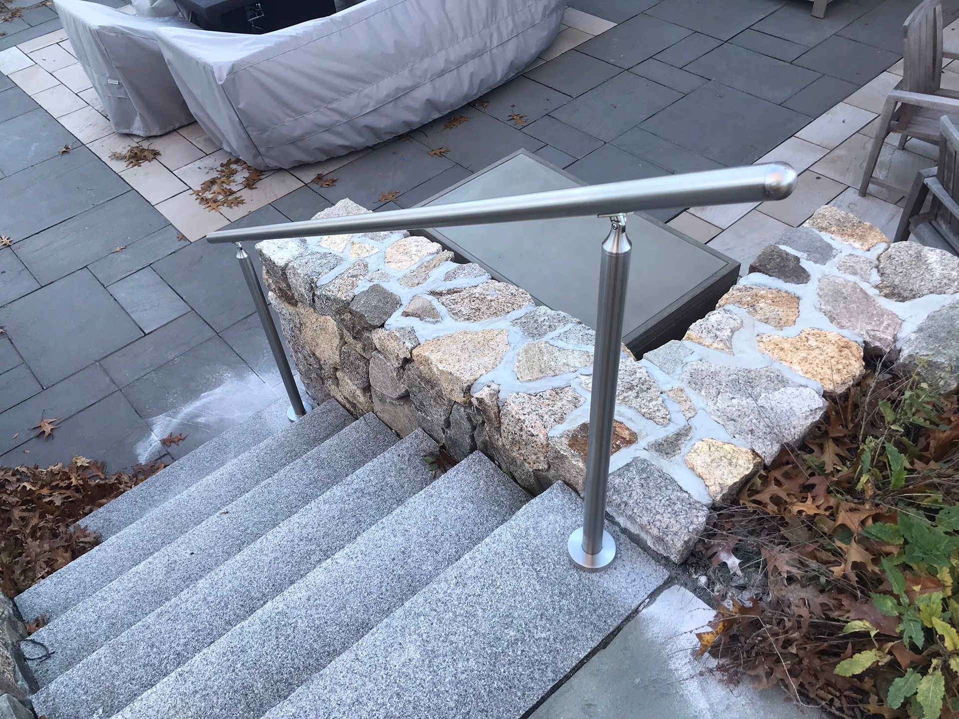 Stone stairway with a silver handrail beside a stone planter and patio, bordered by leaves and plants