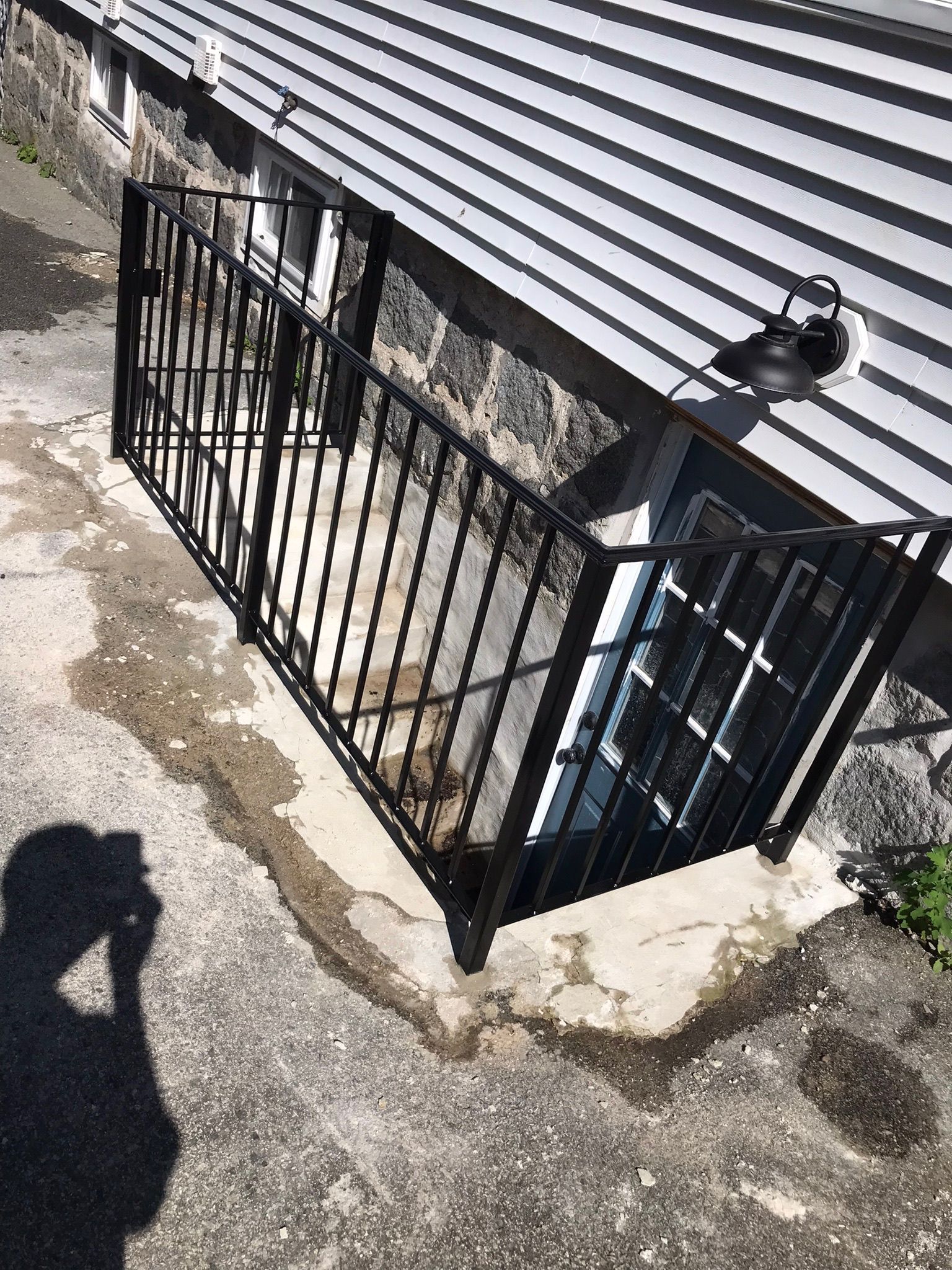 Black metal gate beside a stone house wall and gray roof on a sunlit concrete walkway.