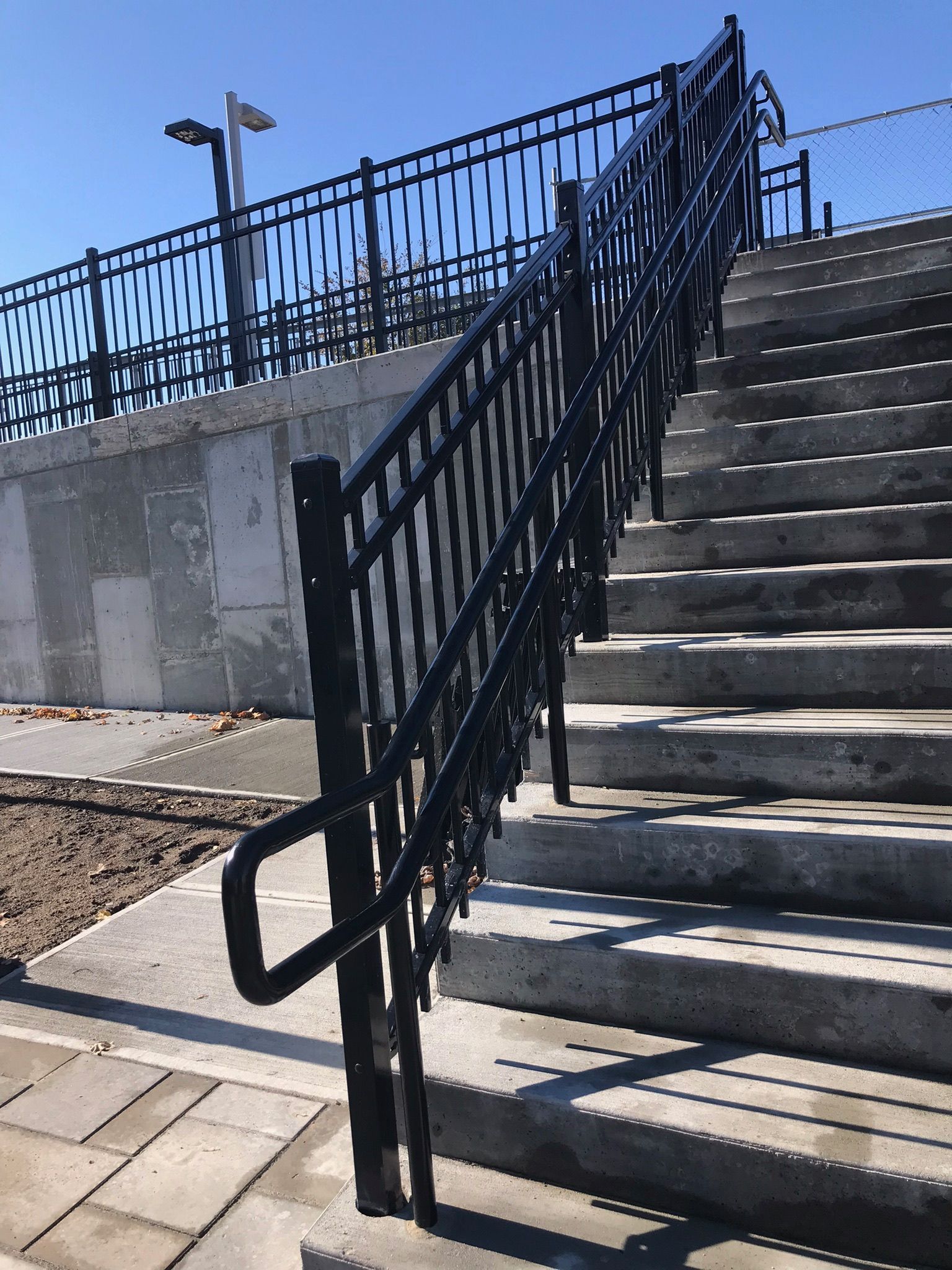 Outdoor stairs with black metal handrails beside a concrete wall and fence under a clear blue sky