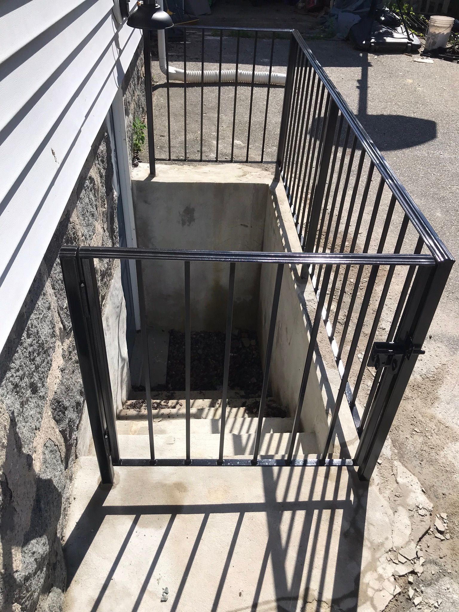 Black metal gate guards basement stairwell beside a stone house and concrete walkway.