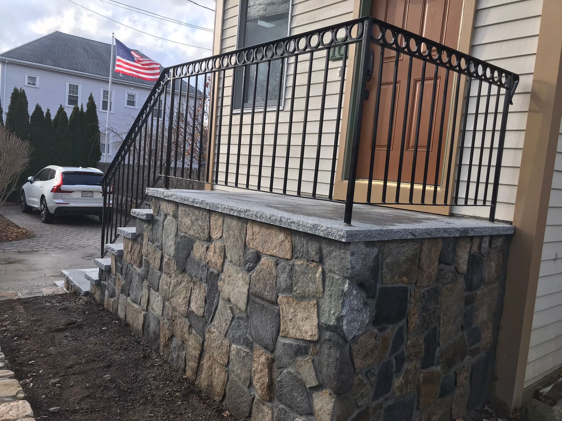 Front porch with stone foundation, black railing, beige siding, and American flag outside a house