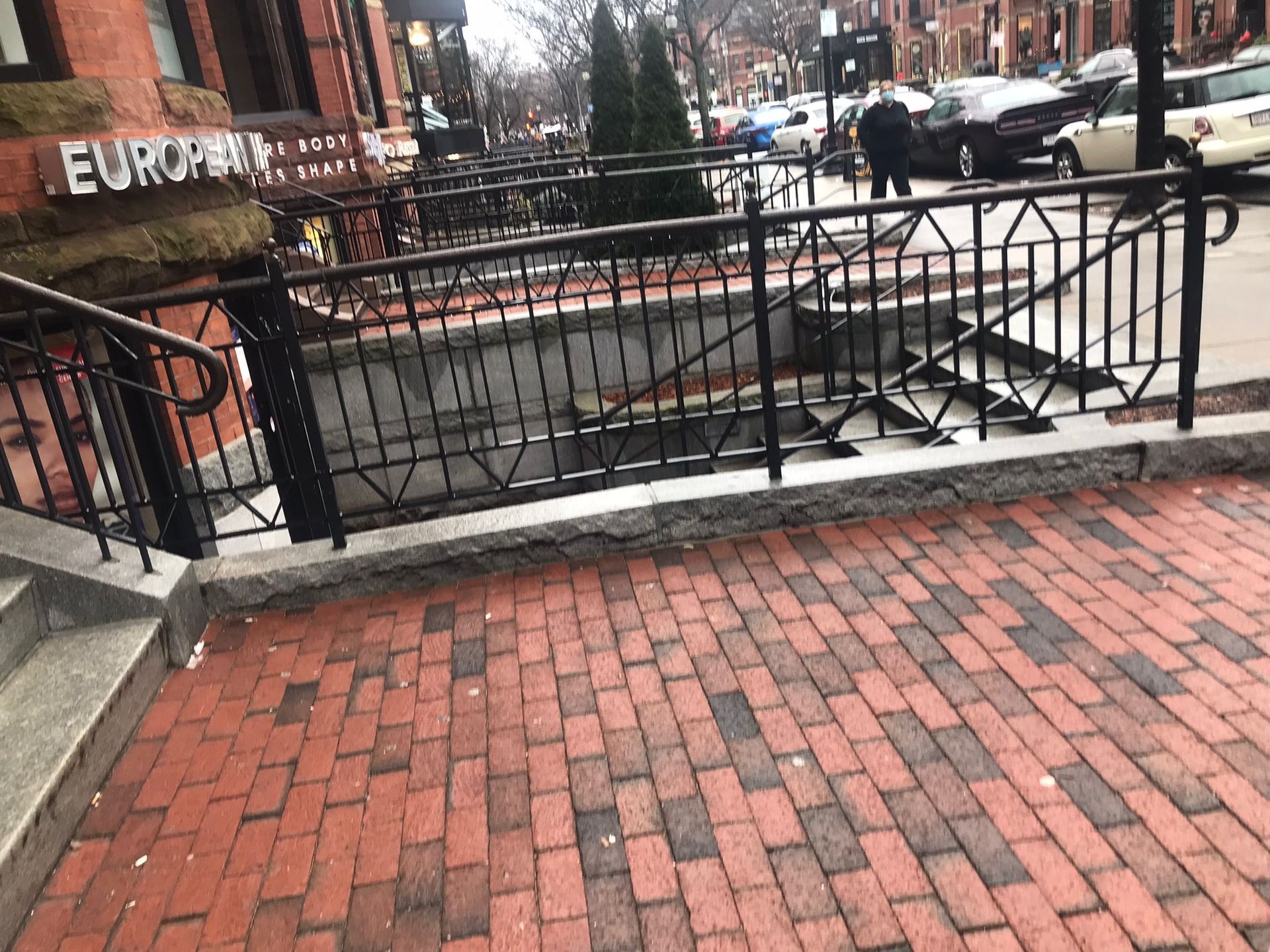Brick sidewalk with a black metal railing overlooking a street and parked cars