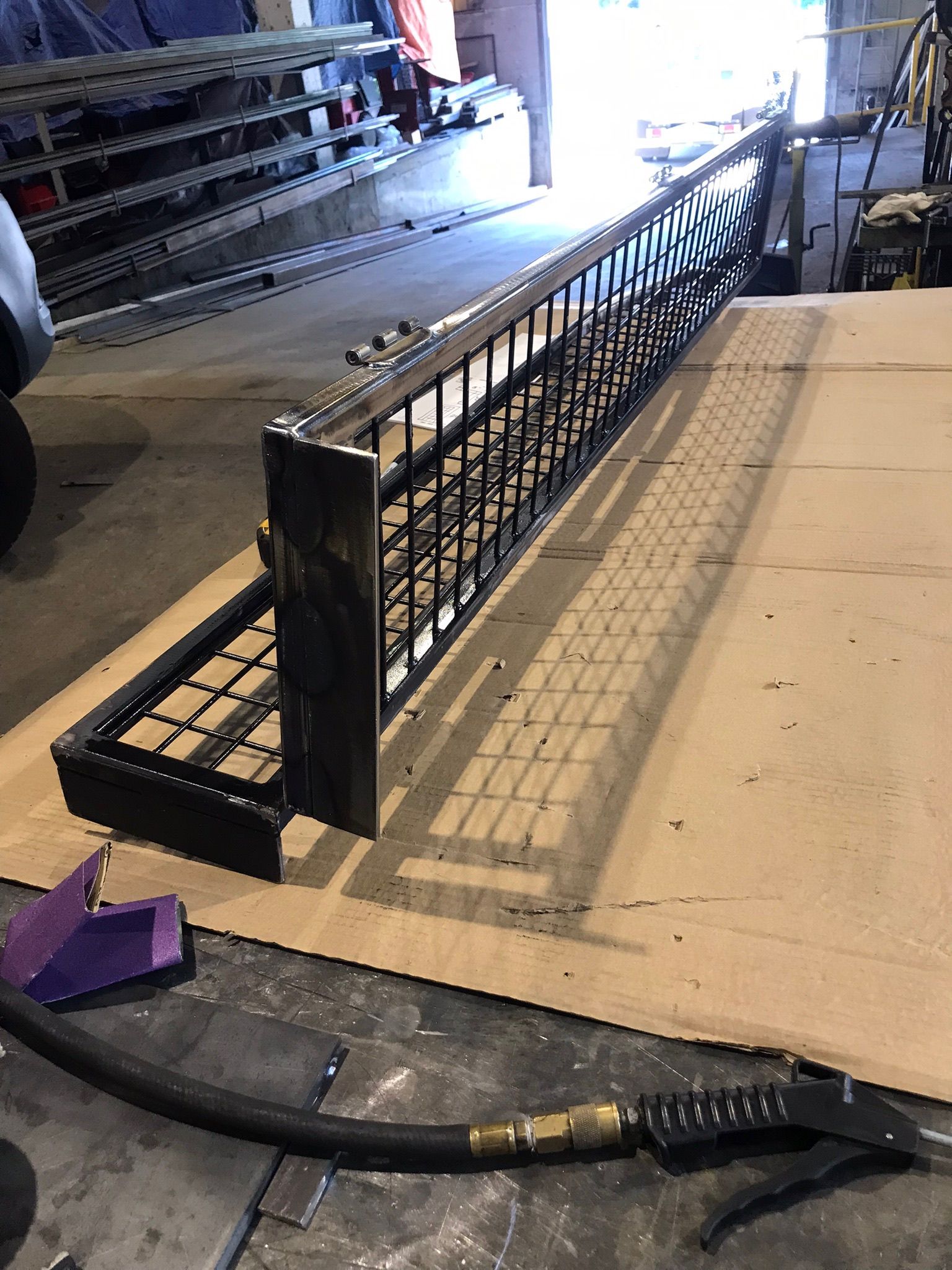 Black metal ramp-like structure in an industrial workshop beside stacked materials and a hose on the floor.