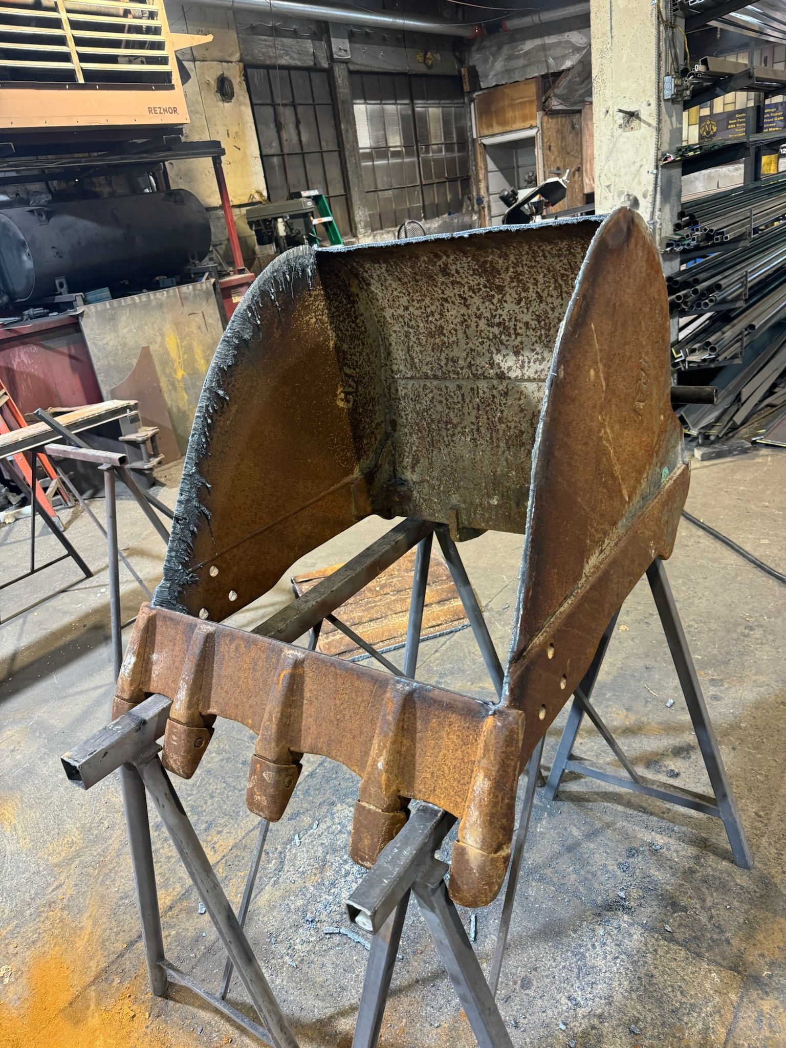 Rusty metal loader bucket in a workshop, propped on stands amid tools and parts.