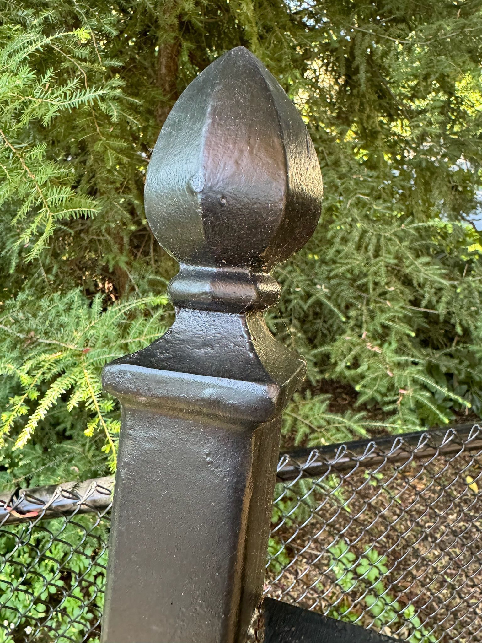 Black decorative fence post finial beside chain-link fence and green foliage