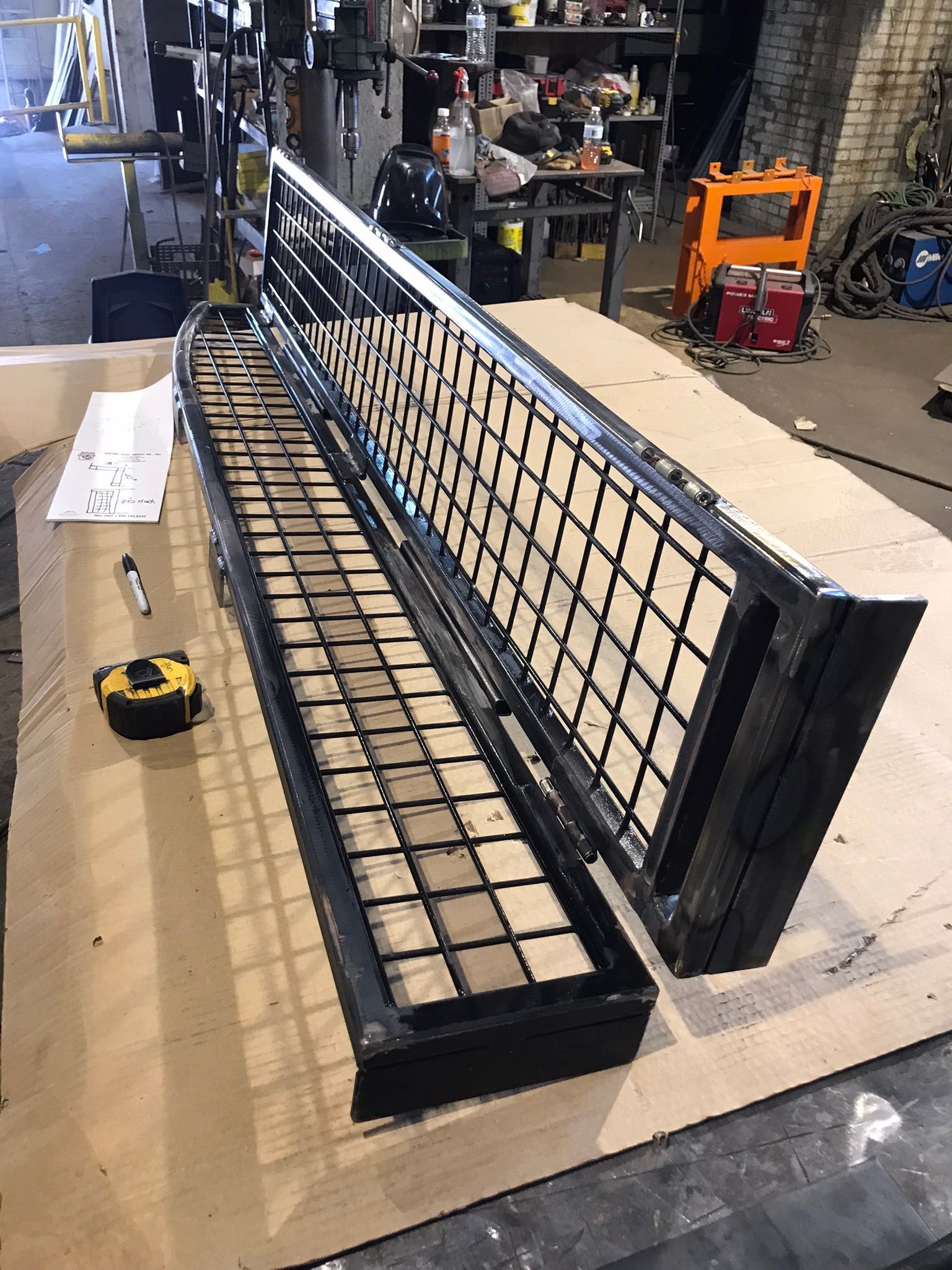 Large black metal mesh gate/door assembly laid on cardboard in a workshop