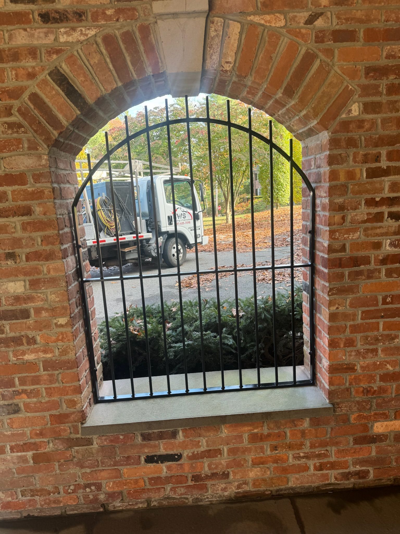 Brick arched window with black iron bars overlooking a garden and parked vehicle outside