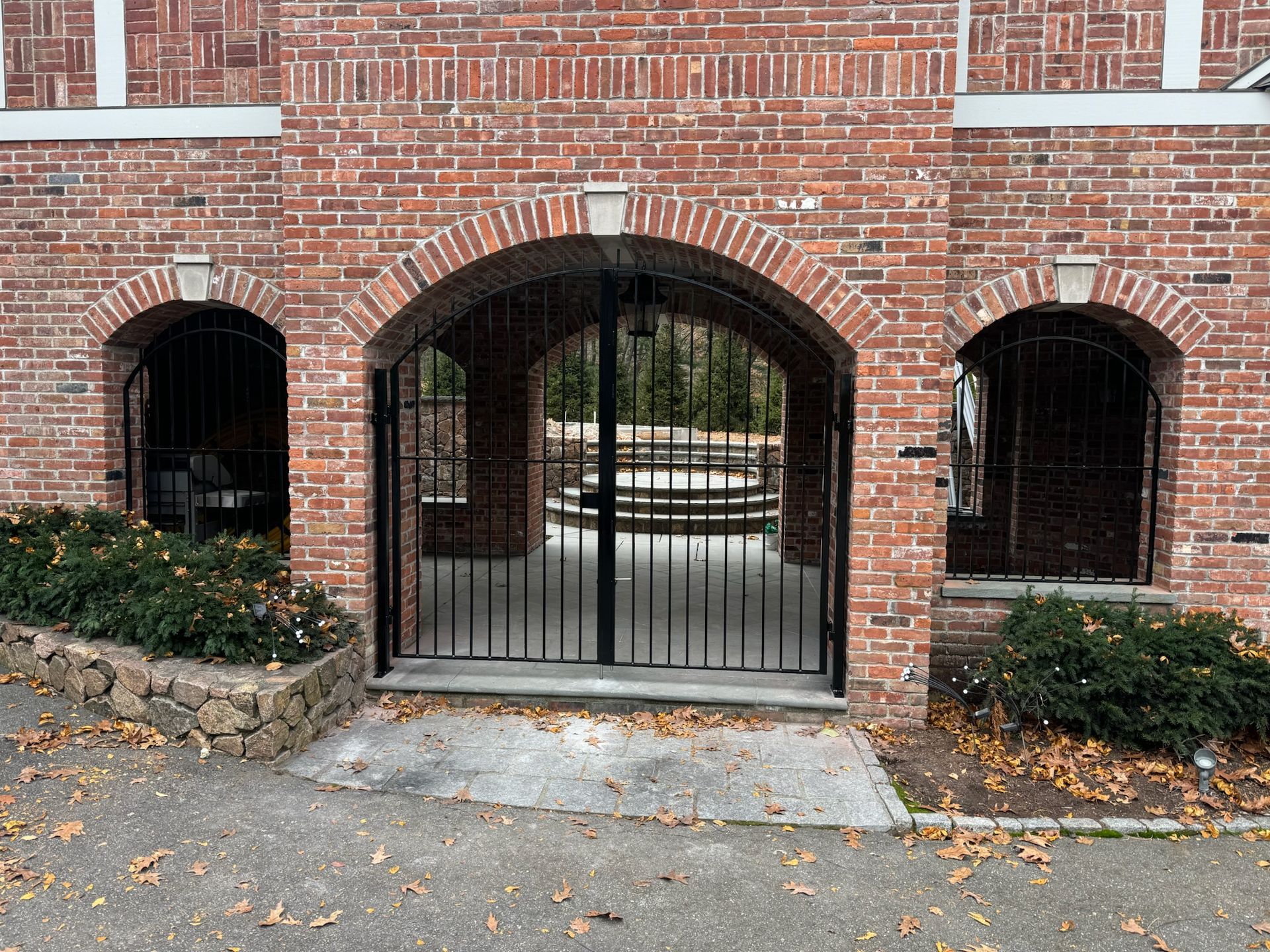 Brick building entrance with a black iron gate and two arched windows, bordered by stone planters.