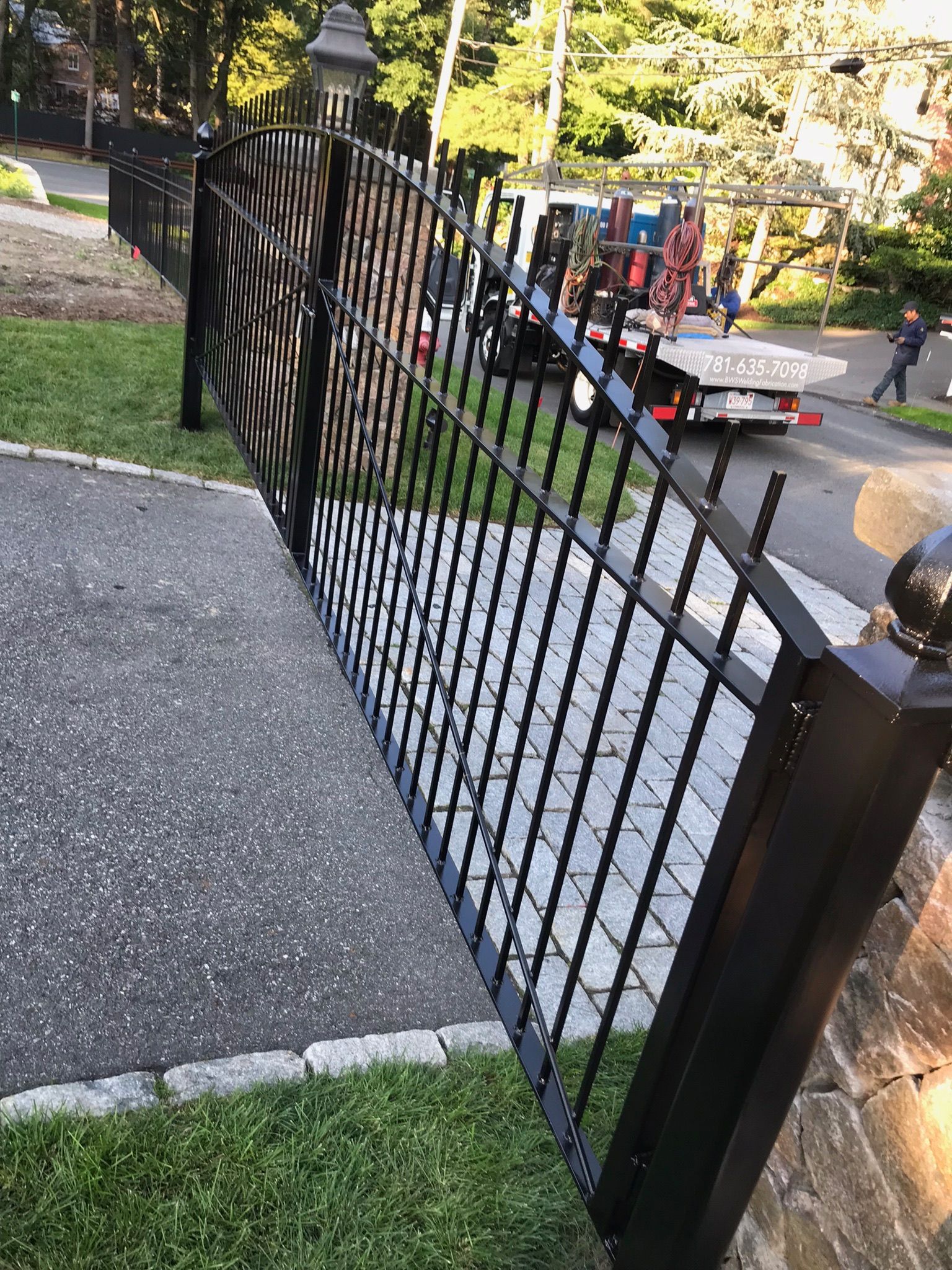 Black metal fence beside a sidewalk, with a street and cars in the background.