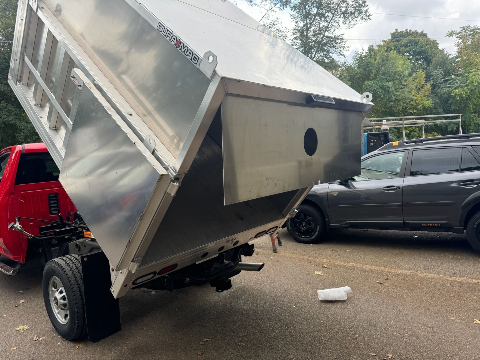 Tilted silver dump trailer parked beside a red truck and black SUV on a street, with debris underneath.