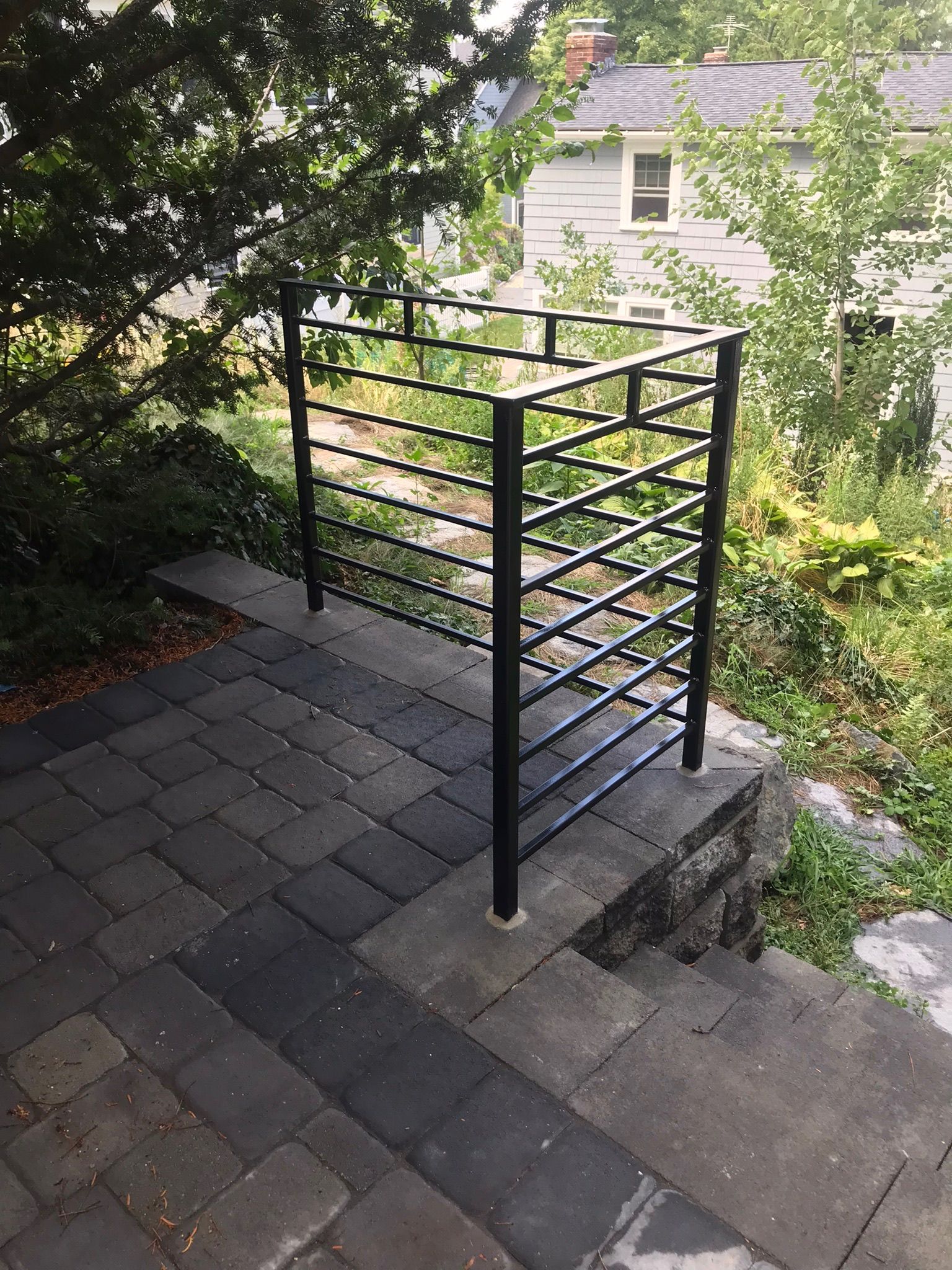 Black metal stair railing beside a gray stone walkway and steps, surrounded by greenery