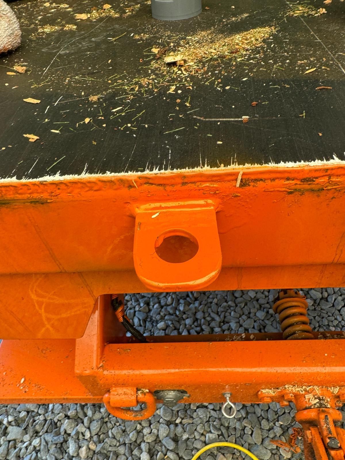Orange metal trailer hitch with black top deck and exposed spring over gravel.