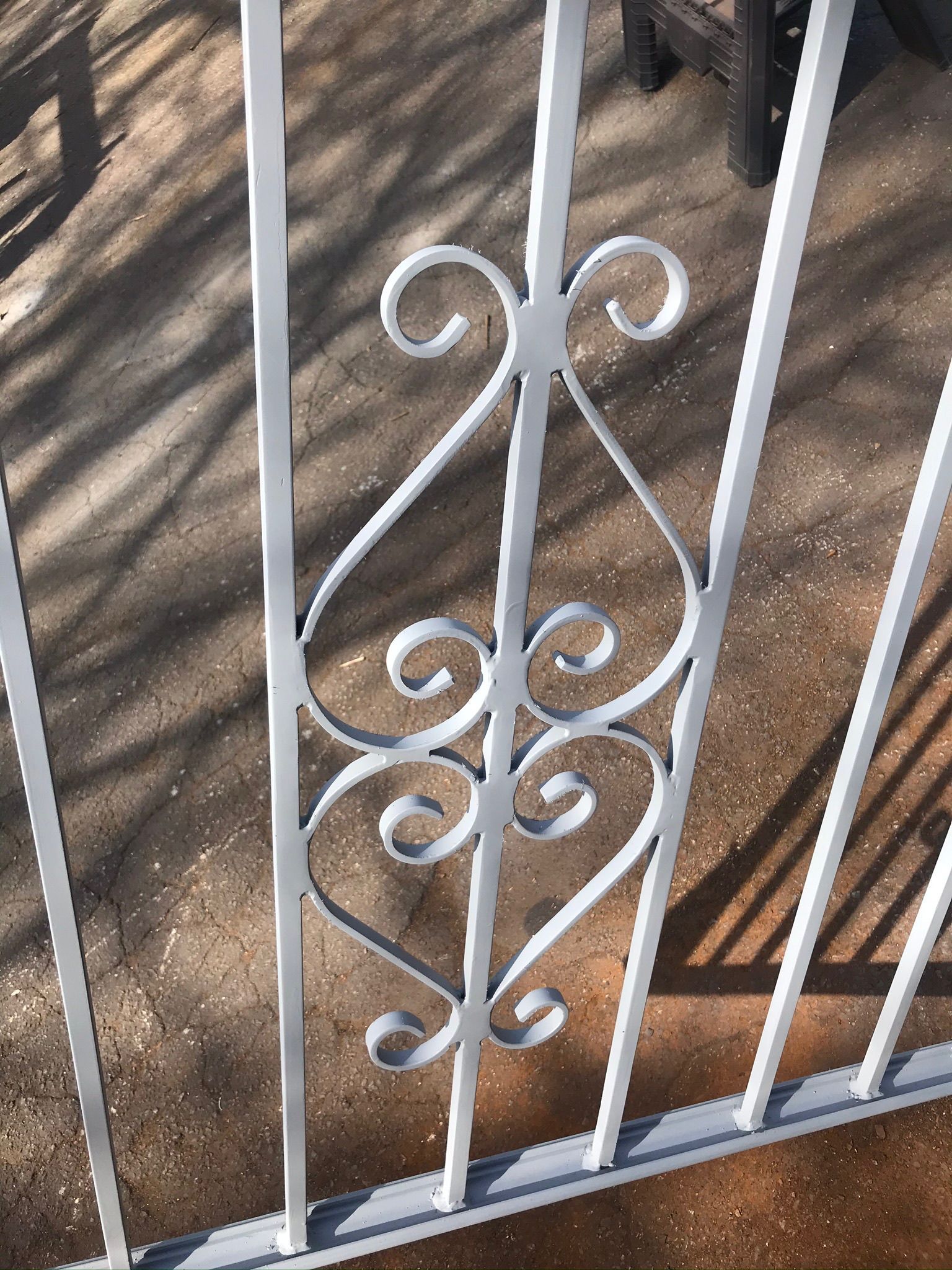 White metal gate with decorative scrollwork in front of a dirt yard and wooden beam.
