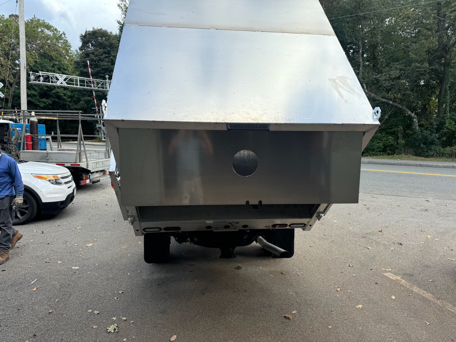 Rear view of a silver truck with a large metal body parked on a roadside near trees and other vehicles