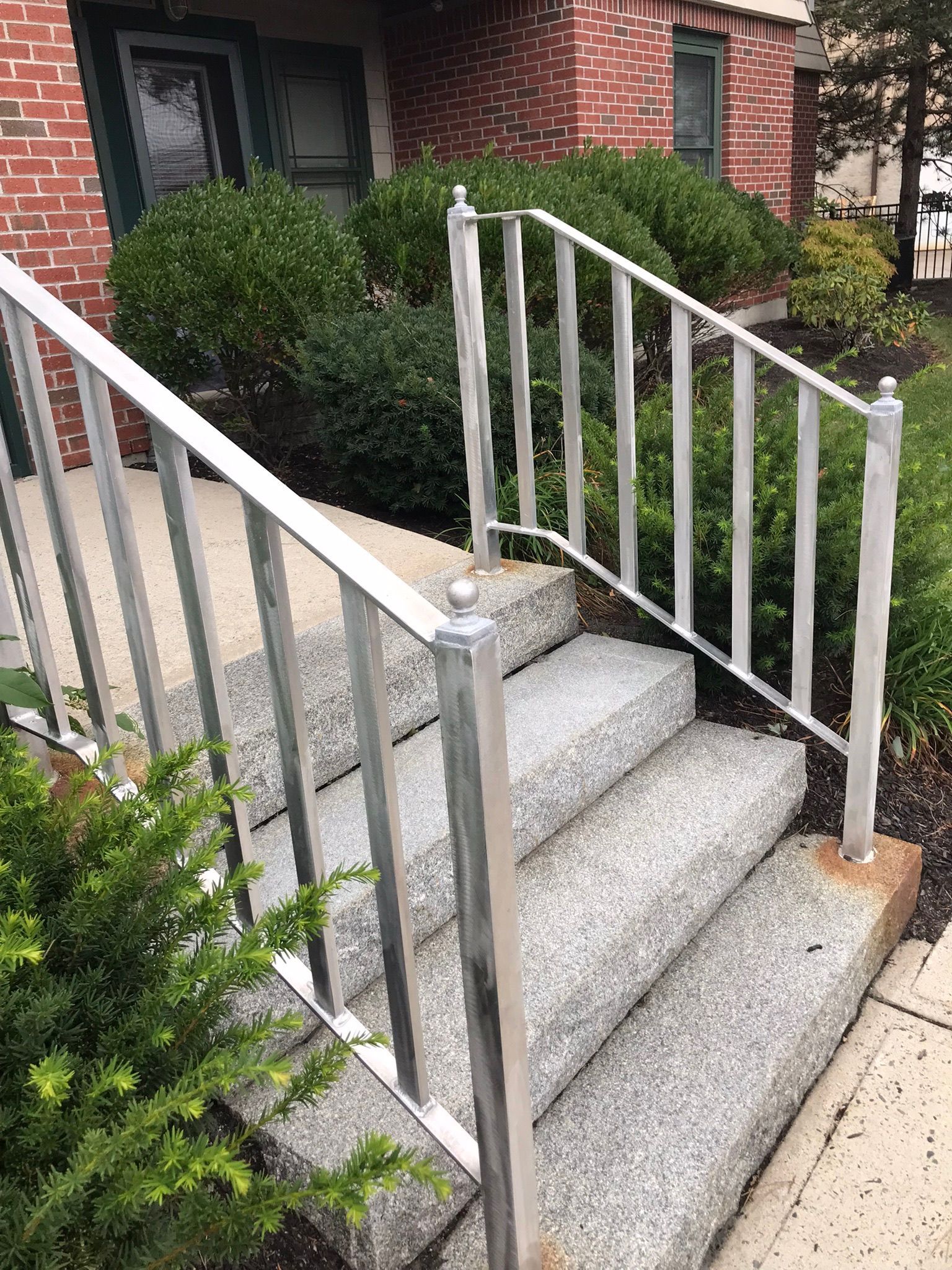 Concrete front steps with white metal railings beside a brick house and shrubs.