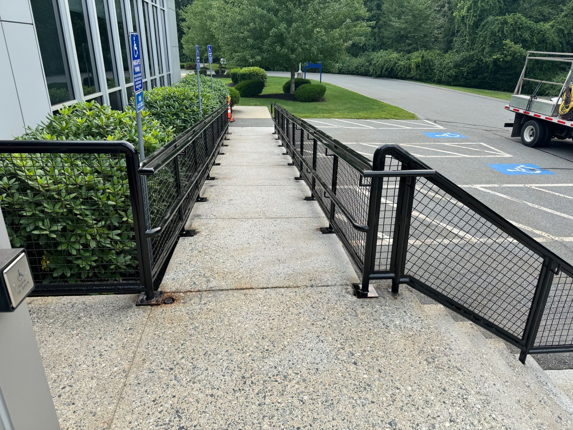 Narrow concrete ramp with black railings beside a sidewalk and parking lot, leading toward a green landscaped area.