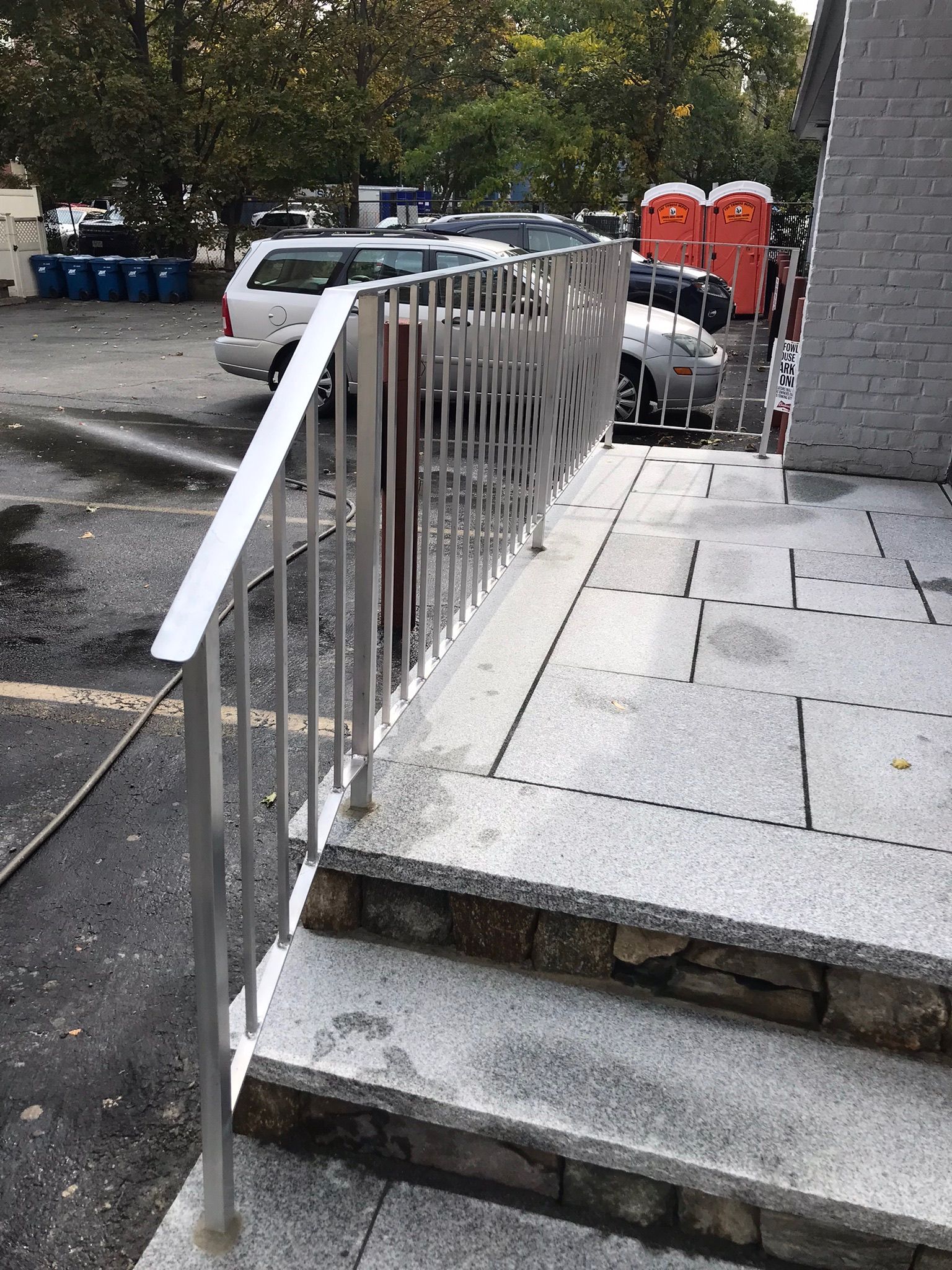 Outdoor stone steps with metal handrail leading to a parking area with cars and red bins
